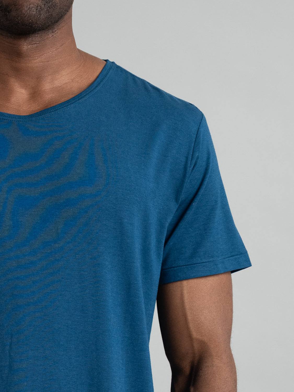Someone wears a Slay Wide Neck Bamboo Blend Tee in blue against a plain gray backdrop. The cropped image shows only the upper torso and part of the right arm. The tee is from ID Menswear.