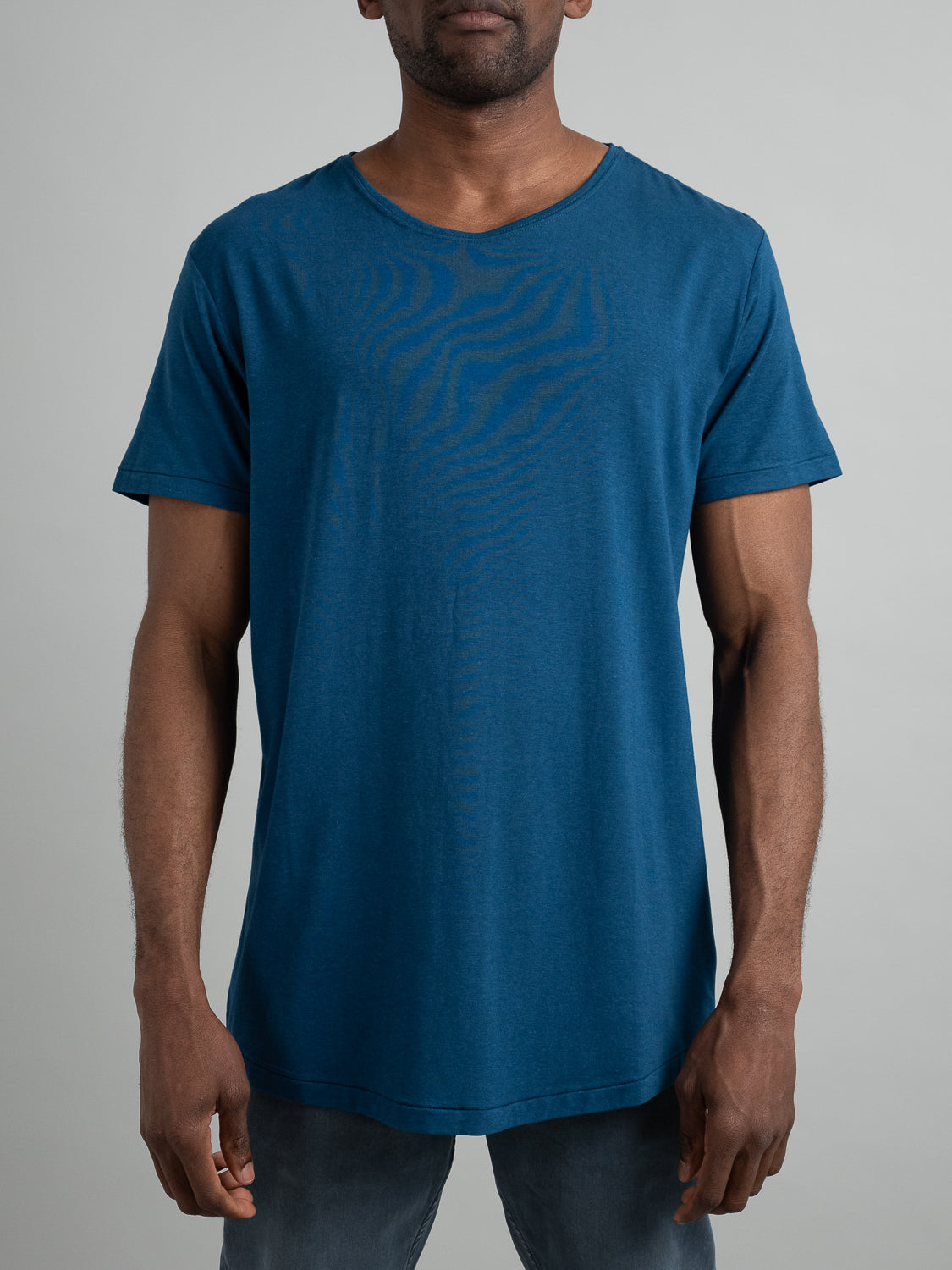 A man in a stylish blue Slay Wide Neck Bamboo Blend Tee from ID Menswear and jeans stands against a plain grey backdrop. The Tee, made of soft bamboo blend fabric, flaunts a subtle textured front pattern. His hands rest casually at his sides.