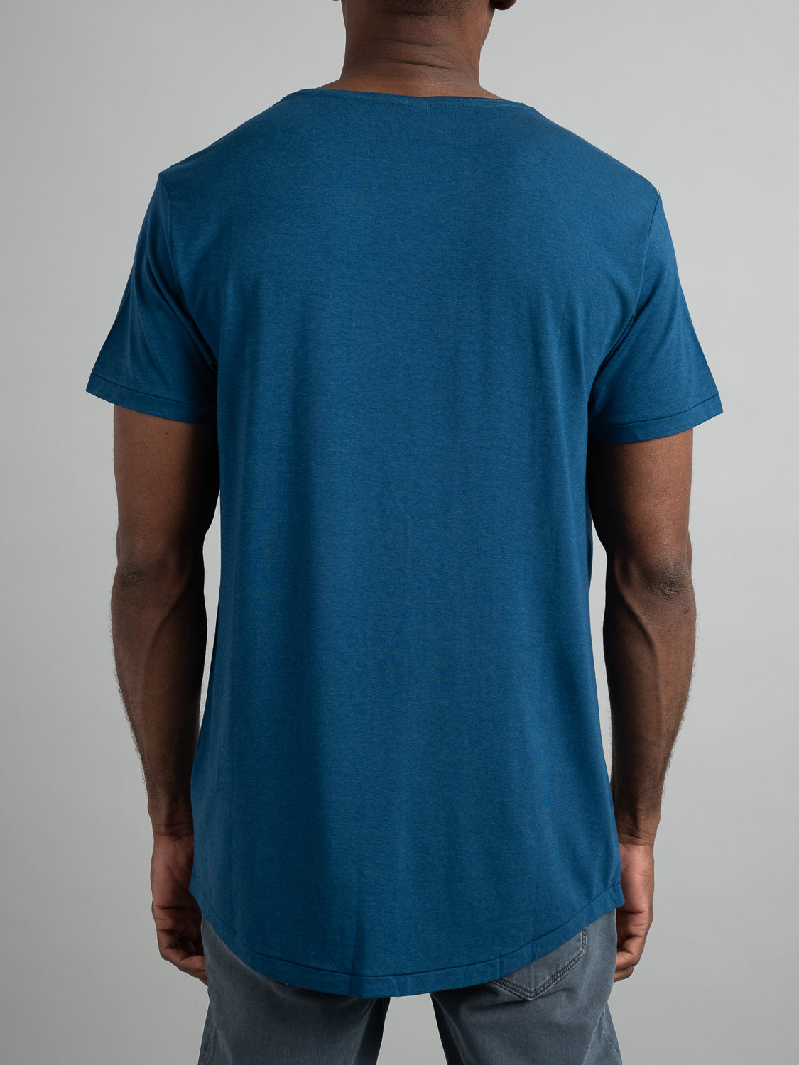 A person stands with their back to the camera, wearing the ID Menswears Slay Wide Neck Bamboo Blend Tee and gray pants, set against a simple, light-colored background. The tee features a straight hem.