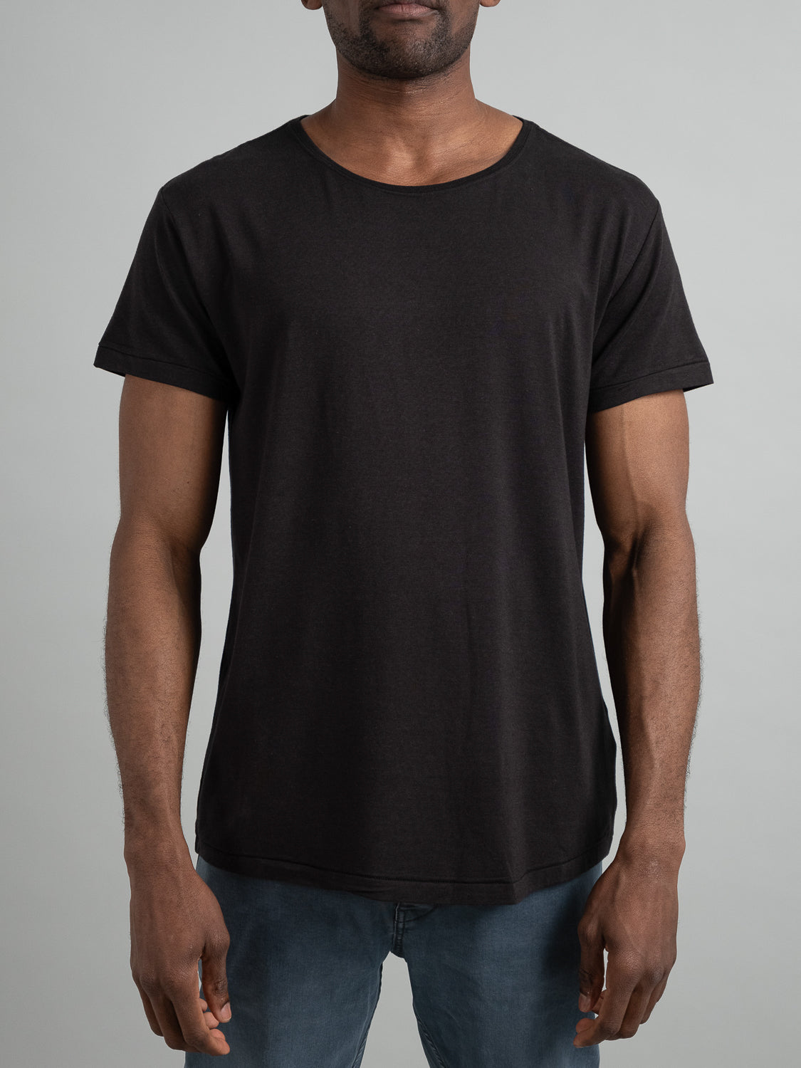 A person stands against a plain light gray background wearing a sleek ID Menswear Slay Wide Neck Bamboo Blend Tee paired with classic blue jeans. The focus is on the torso, with arms hanging by the sides.