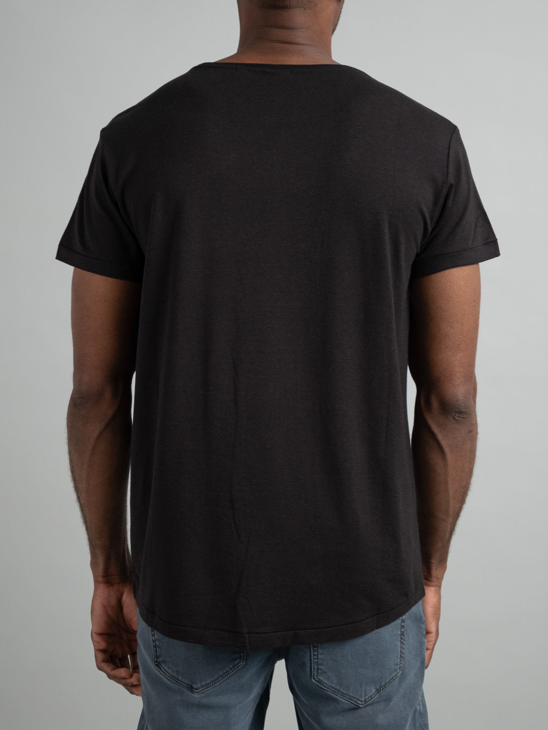 A person is wearing an ID Menswear Slay Wide Neck Bamboo Blend Tee in plain black with blue jeans, standing with their back to the camera against a neutral background.