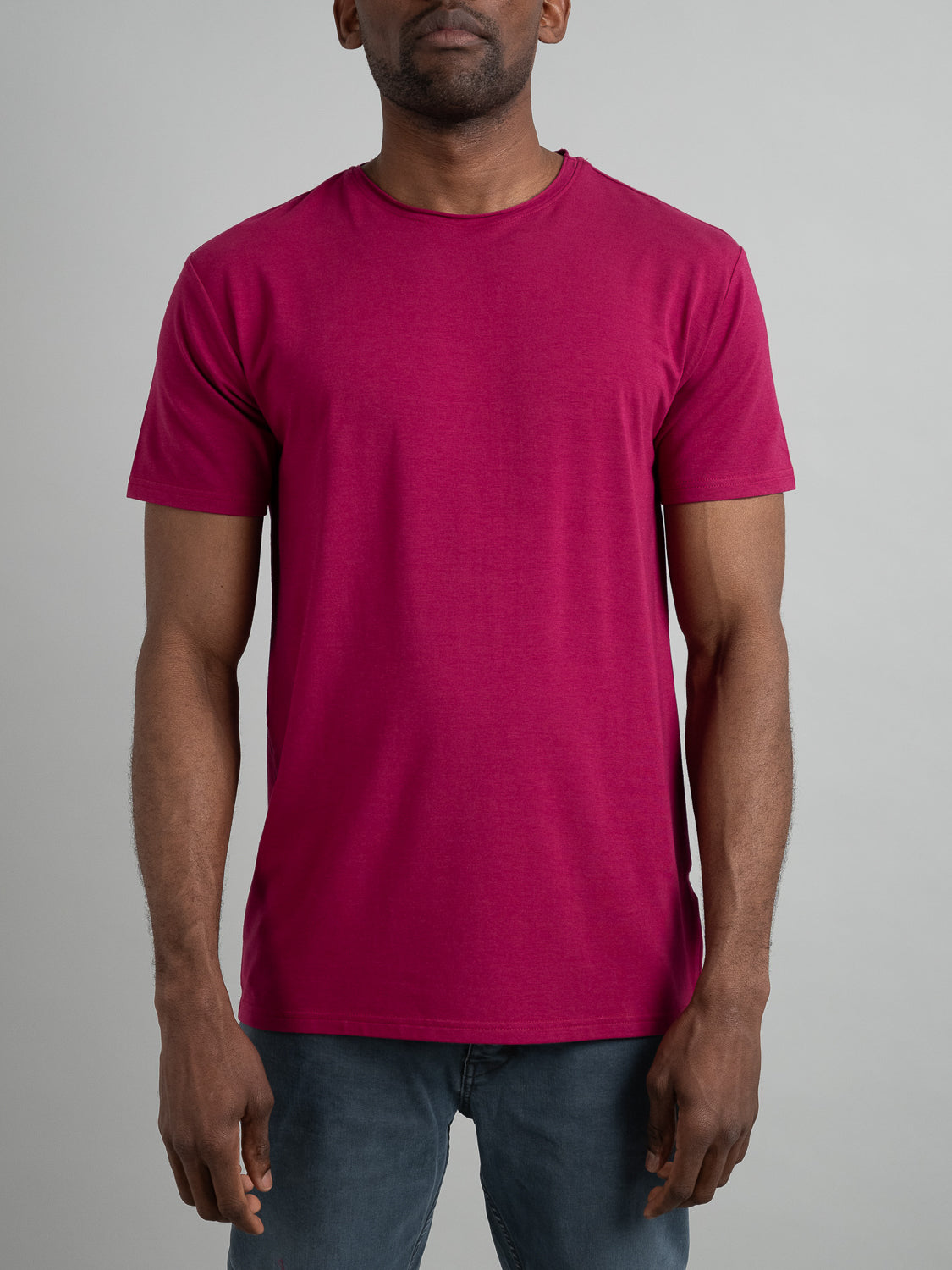 A man stands confidently against a gray backdrop, wearing the Bello Bamboo Blend Rolled Neck Tee by ID Menswear—a vibrant pink t-shirt made with organic fabric—paired with gray pants, showcasing both stylish design and sustainable fashion.
