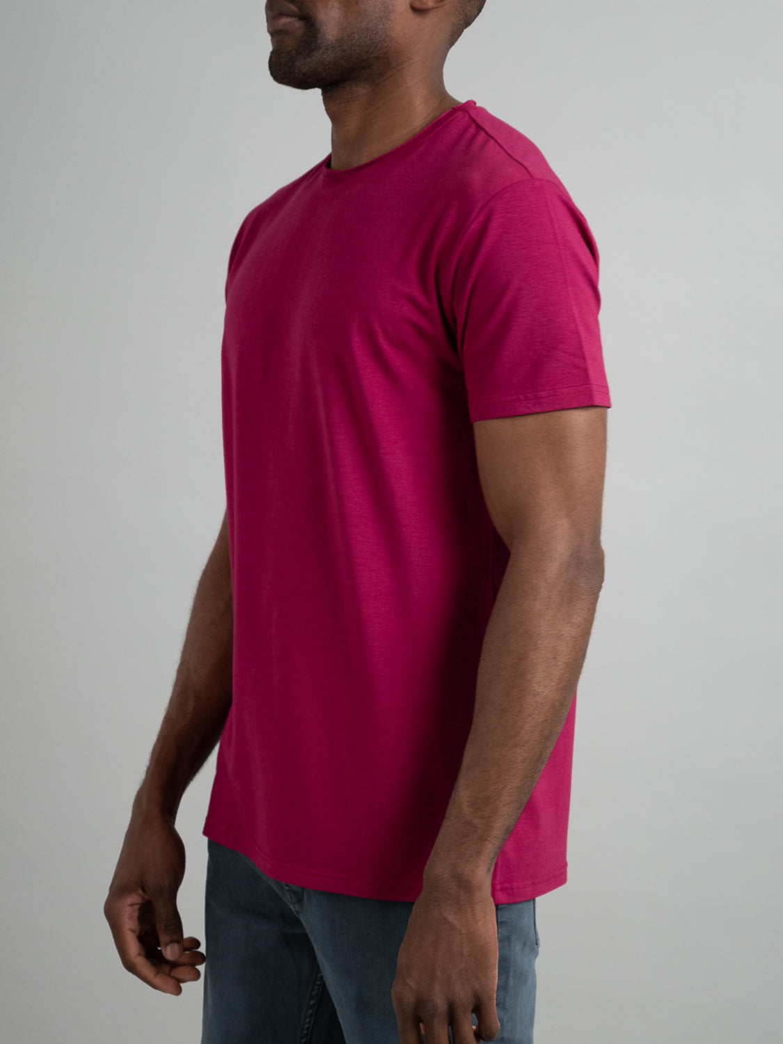 A person in a Bello Bamboo Blend Rolled Neck Tee by ID Menswear, featuring bright magenta color and organic cotton, paired with blue jeans, stands against a neutral backdrop. The image emphasizes their sustainable fashion choice through an upper body side profile view.