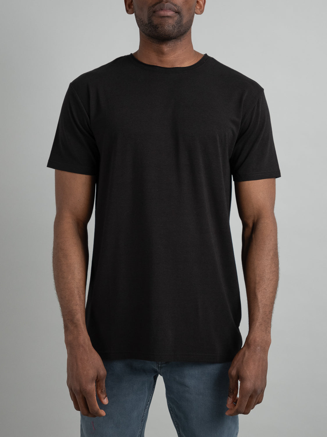 A person stands against a gray background, wearing the ID Menswear Bello Bamboo Blend Rolled Neck Tee and blue jeans. This short-sleeved tee, with its casual fit, highlights a sustainable clothing choice while arms are relaxed at the sides.