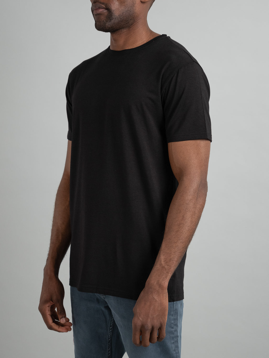 A man in a Bello Bamboo Blend Rolled Neck Tee from ID Menswear and blue jeans stands against a neutral background, his hands relaxed by his sides.