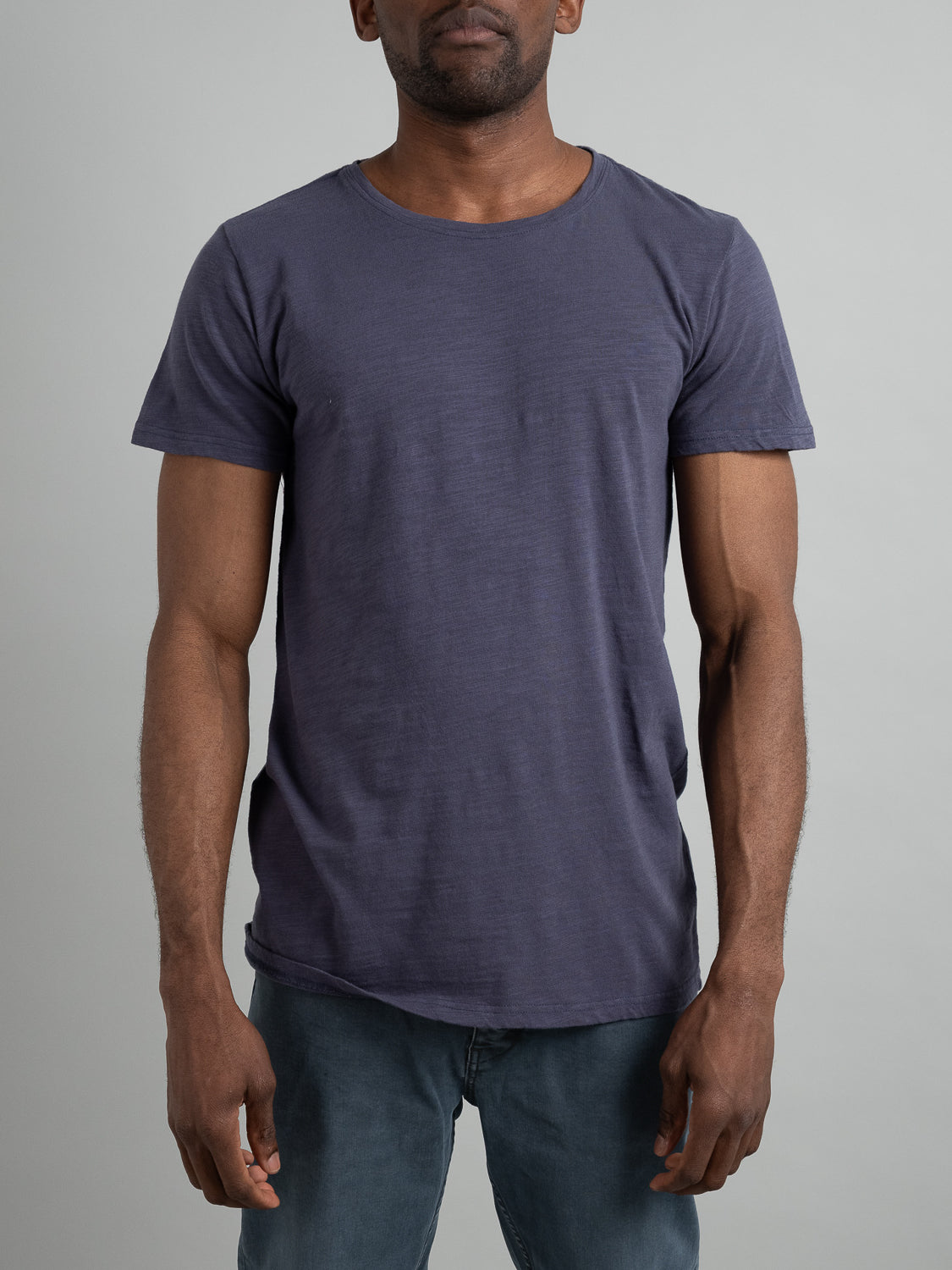 A person wearing a Mini Flip Slub Yarn Tee by ID Menswear and faded blue jeans stands against a neutral gray background. The image highlights the upper body, projecting a relaxed and casual look perfect for everyday wear.