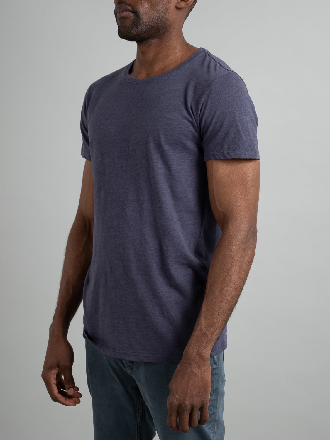 Wearing ID Menswears Mini Flip Slub Yarn Tee, a plain, short-sleeved, grayish-blue T-shirt, paired with dark jeans, a person stands against a light gray backdrop. They face slightly left, capturing a casual vibe with relaxed arms.