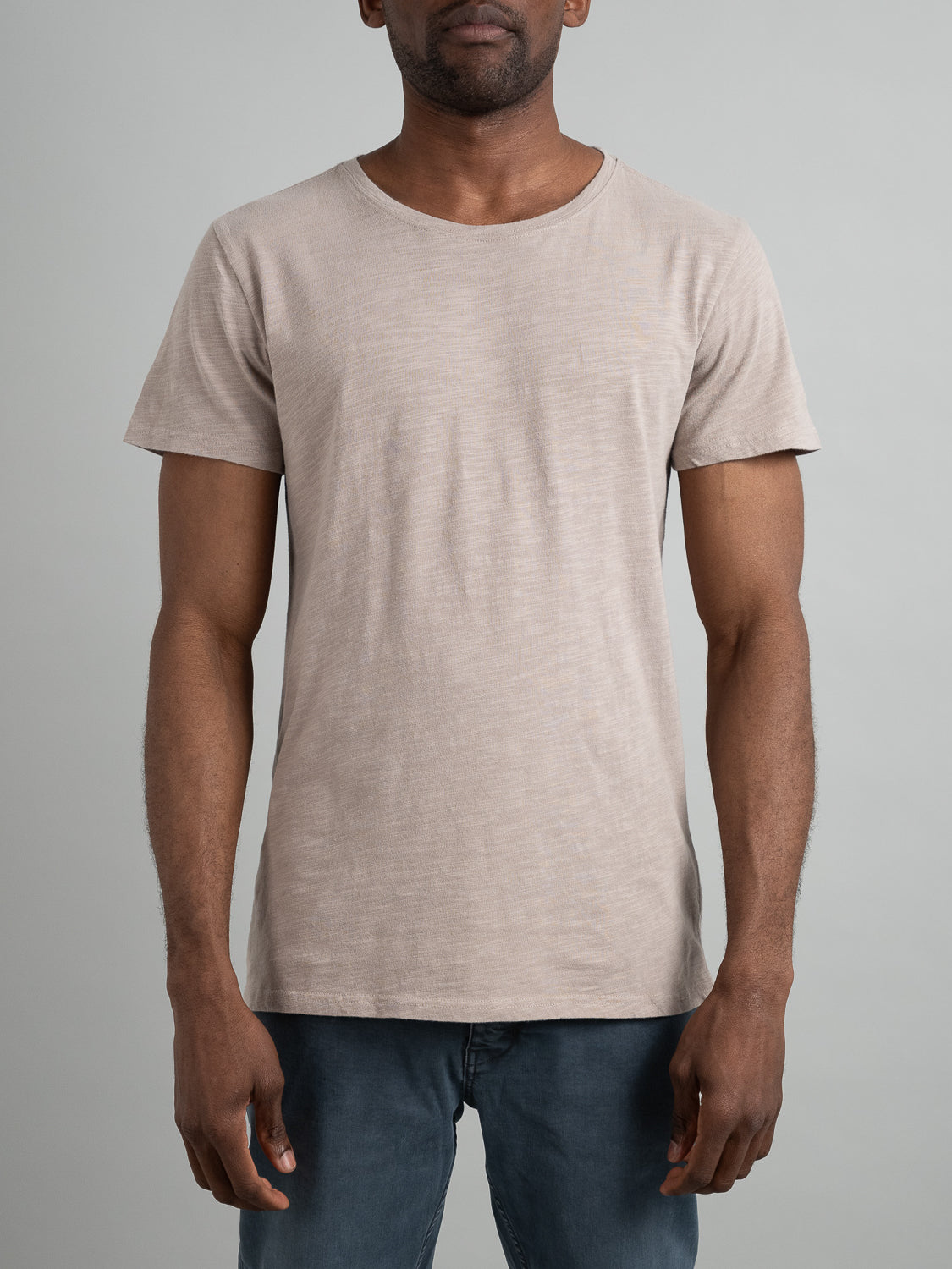 A person in the Mini Flip Slub Yarn Tee by ID Menswear paired with blue jeans stands against a gray backdrop. The casual beige tee, featuring a round neckline and short sleeves, creates an effortless look as the persons arms rest calmly at their sides.