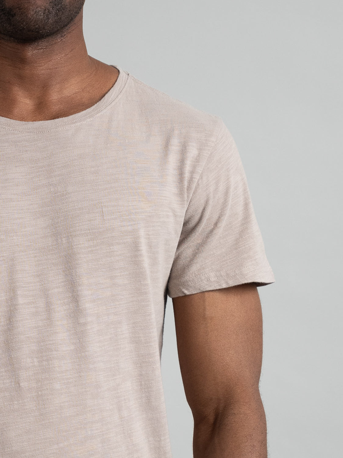 A close-up showcases the Mini Flip Slub Yarn Tee by ID Menswear. The light beige slub cotton tee stands out against a plain gray background, focusing on the upper torso and arm for a casual, everyday look. Part of the face and left side are out of frame.
