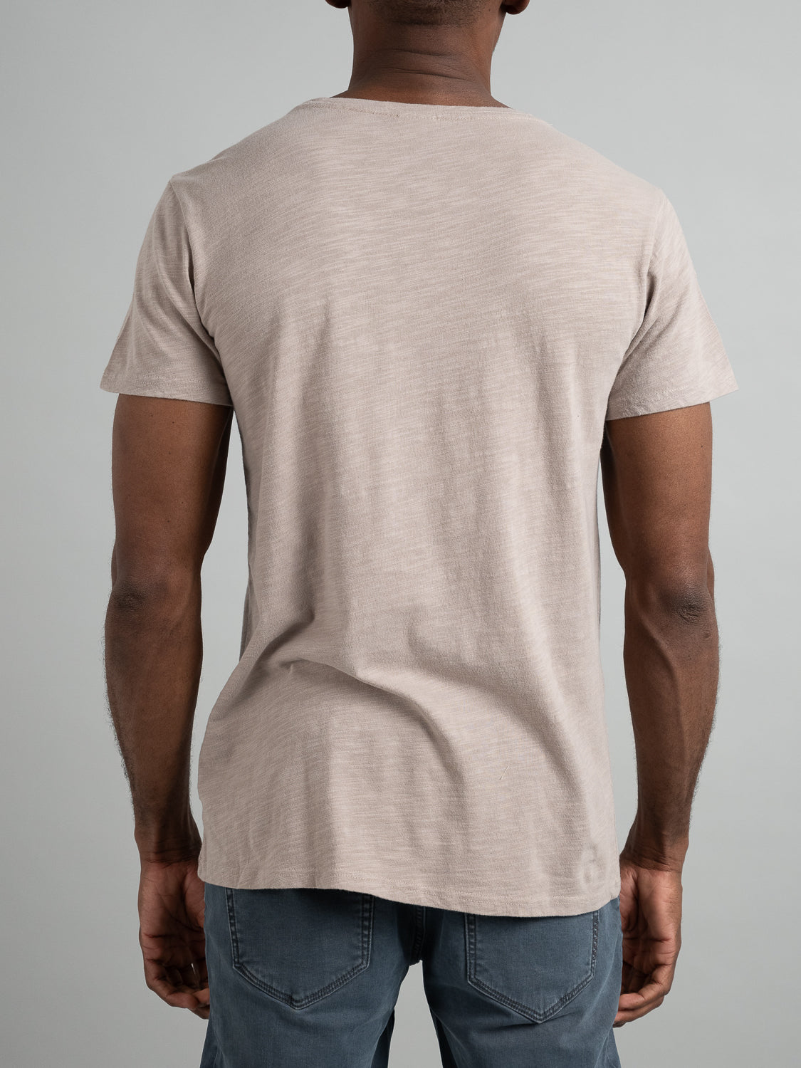 A person is seen from the back, exhibiting a casual look with an ID Menswear Mini Flip Slub Yarn Tee in light beige and blue jeans, set against a plain gray background.