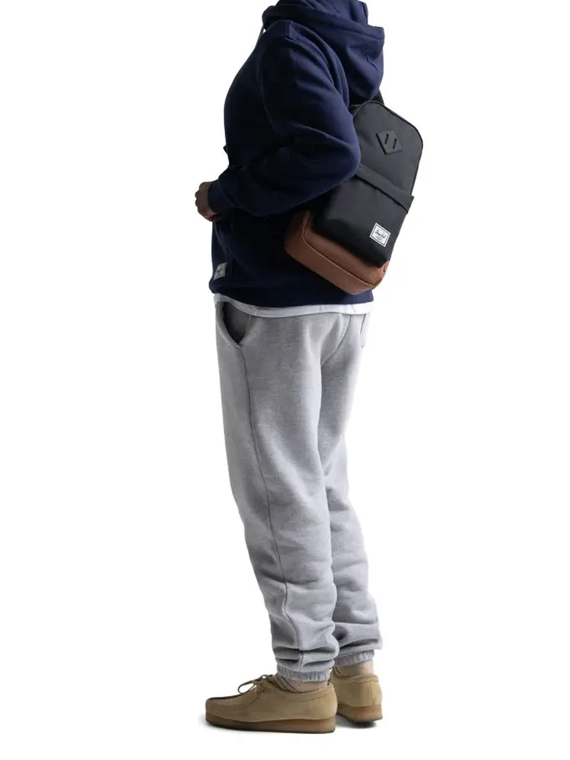 A person in a navy hoodie, gray sweatpants, and tan shoes stands in profile against a white background, carrying the oversized Herschel Heritage Shoulder Bag.
