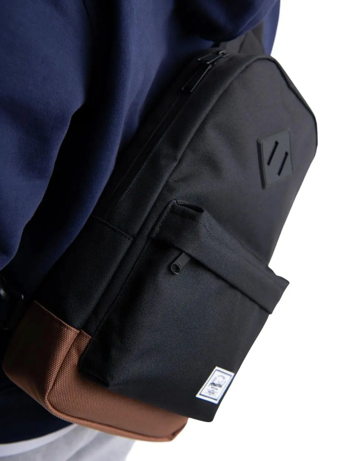 A person wears a Herschel Heritage Shoulder Bag in black and brown over a navy blue hoodie. The bag features a front pocket with a white Herschel logo tag and a geometric patch on the main compartment.