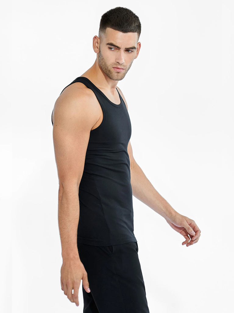 A man with short dark hair wears a Bread & Boxers 2 Pack Tank Top Cotton Stretch and black pants. He looks to his left while walking against a plain white background.