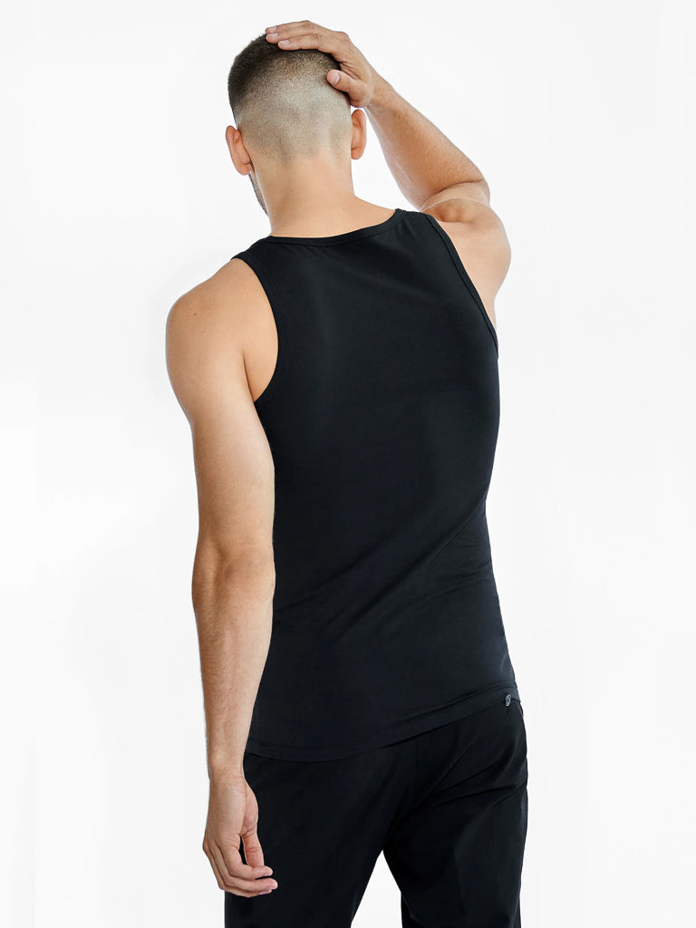 A man with a shaved head wears the Bread & Boxers 2 Pack Tank Top Cotton Stretch in black, paired with matching pants. Facing away, his hand rests on his head against a white background, highlighting his minimalistic style.