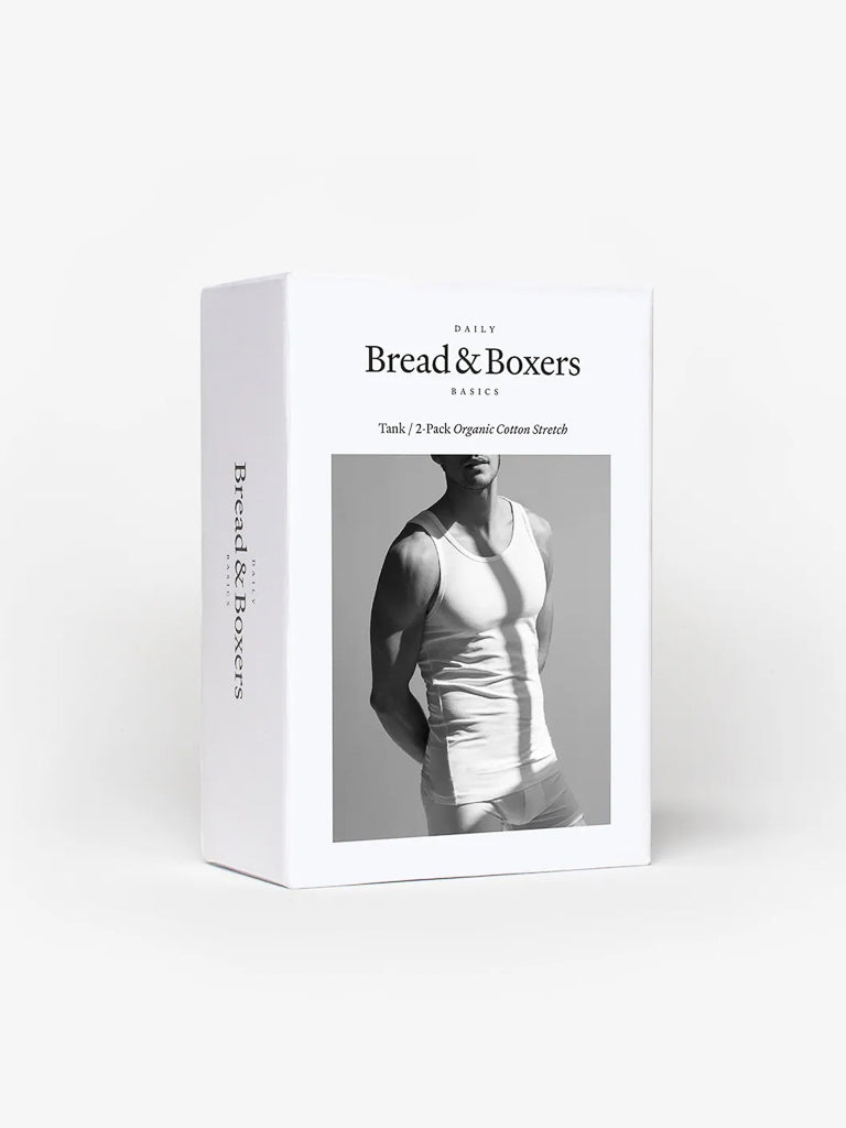 A white box displays a person in a white tank top. Its by Bread & Boxer, featuring their 2-Pack Tank Top in organic stretch cotton. Ideal as a base layer or standalone piece, these premium tanks offer both comfort and style.