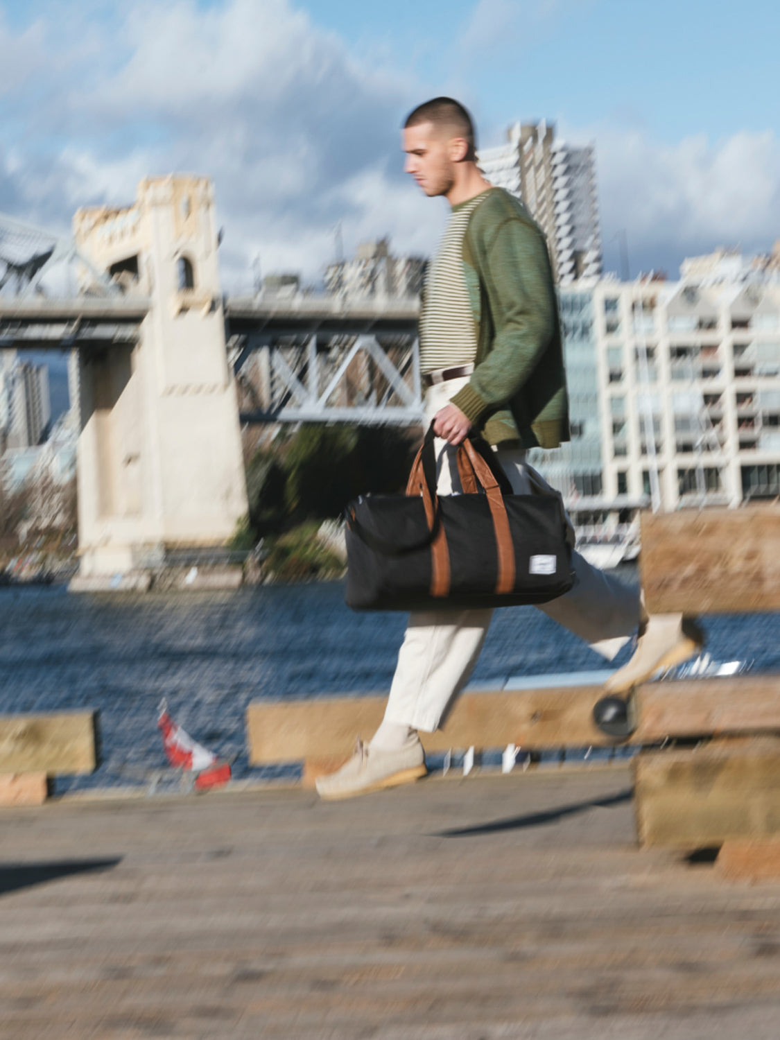 A man walks along a dock carrying a black Herschel Novel Weekender Duffle Bag with a shoe compartment. He wears light pants, a striped shirt, and a green jacket, with a bridge, water, and city buildings under a partly cloudy sky behind him.