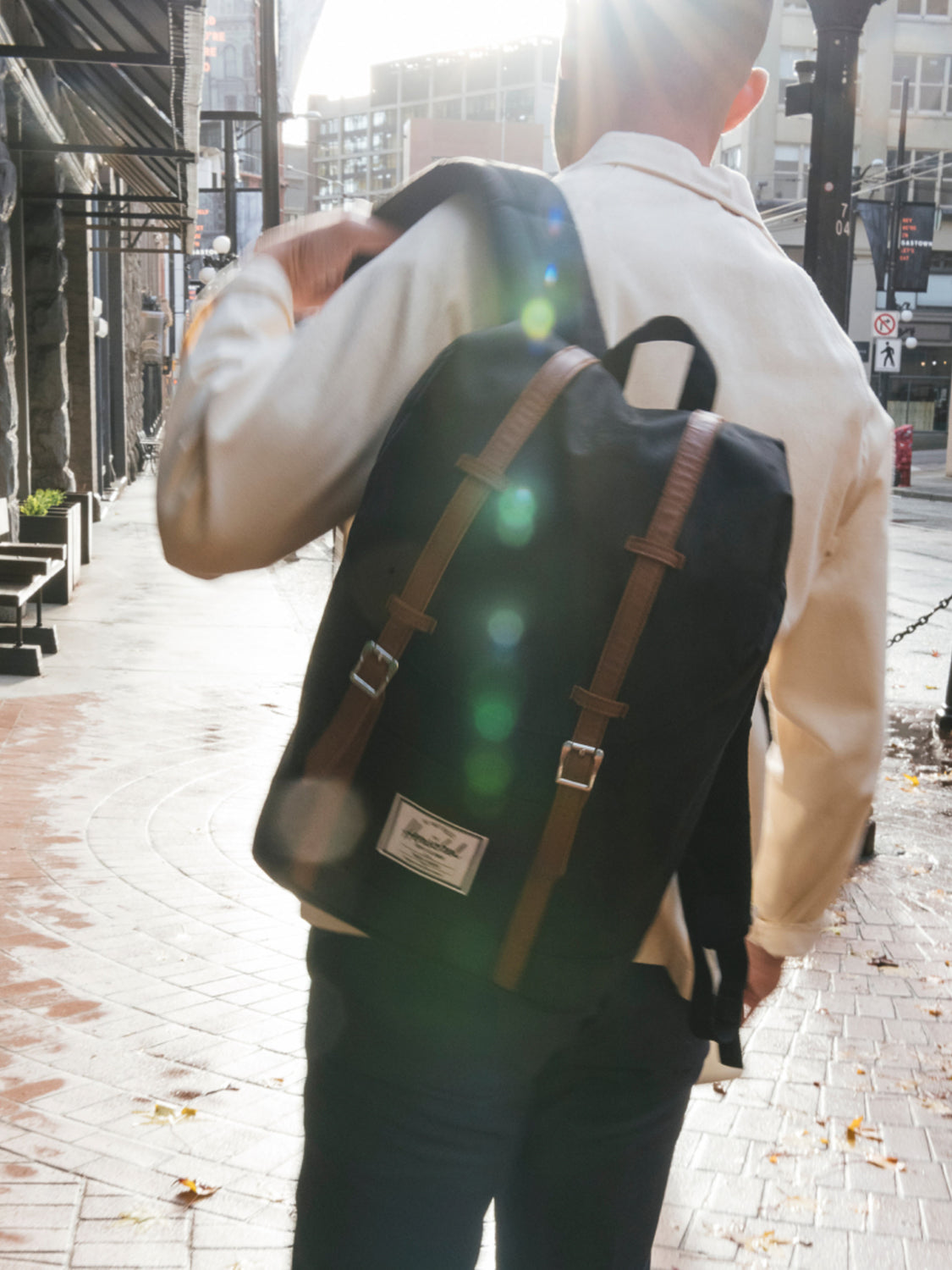 A person with short hair in a light jacket walks on a sunny city sidewalk, carrying a Herschel Retreat Backpack with brown straps over one shoulder.