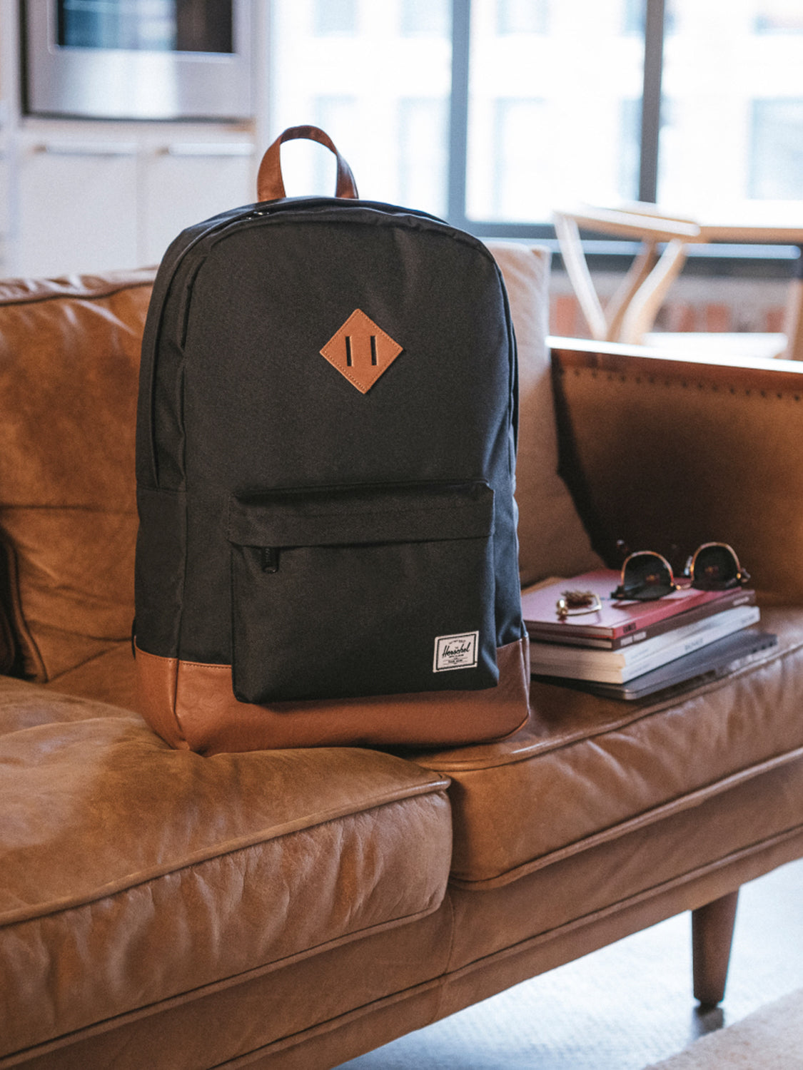 A Herschel Heritage Backpack with a laptop sleeve rests on a tan leather couch beside books, magazines, and sunglasses in a bright living room—offering classic mountaineering style with modern urban flair.