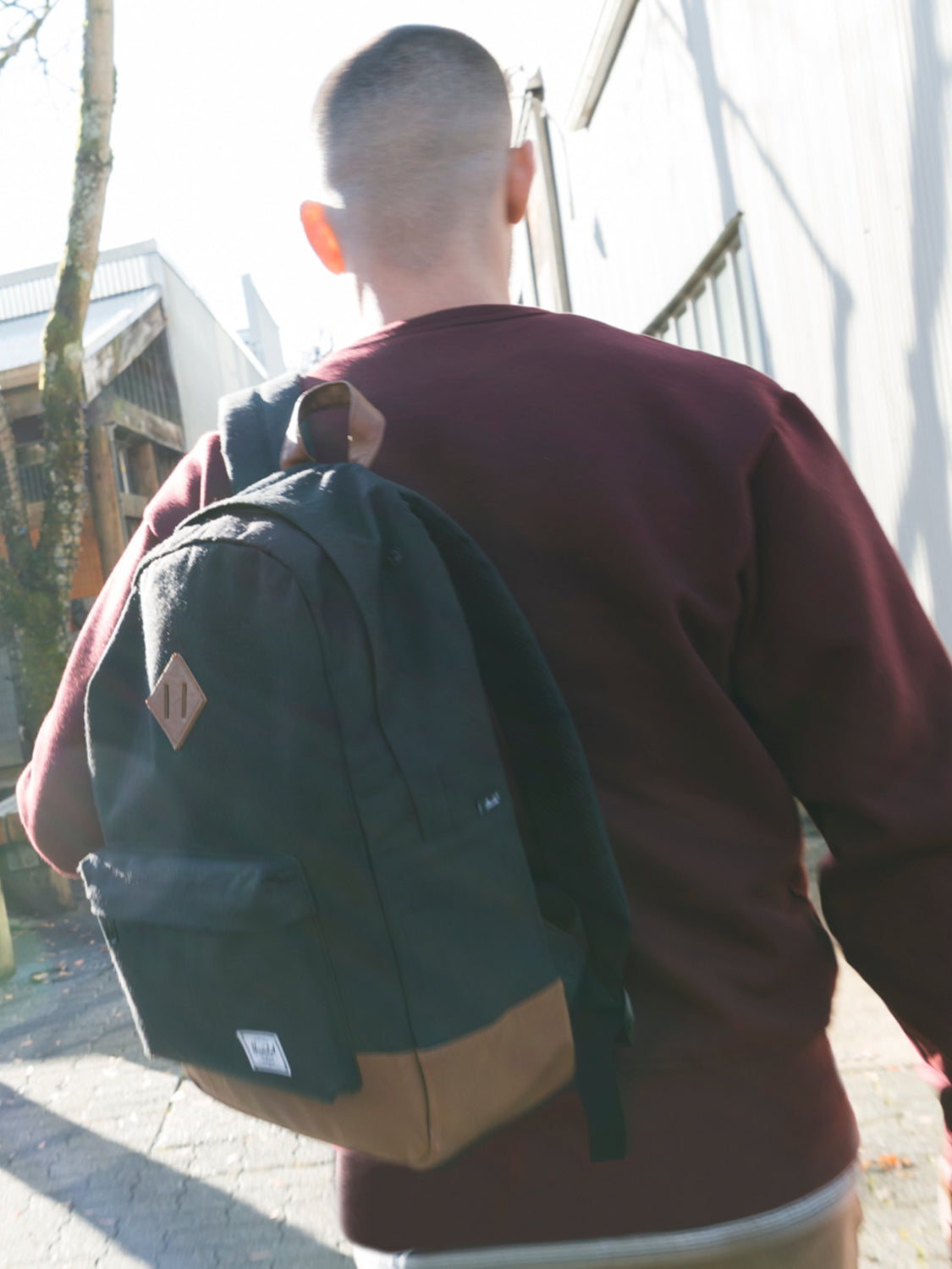 A person with short hair, wearing a maroon sweater and carrying the Herschel Heritage Backpack with a laptop sleeve, walks outdoors on a sunny day beside light-colored buildings.