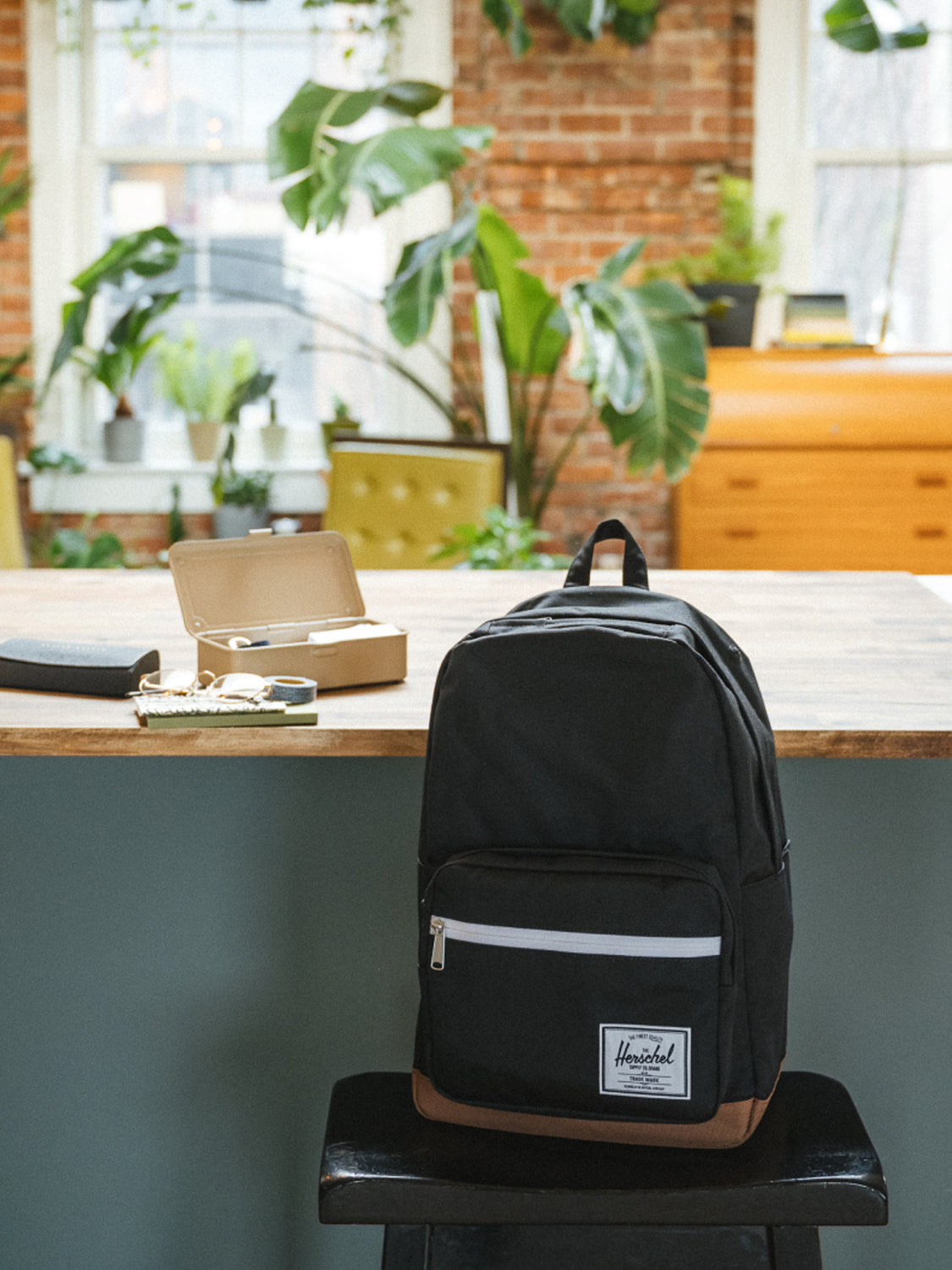 A black and brown Herschel Pop Quiz Backpack with a laptop sleeve rests on a stool next to a notebook, pen, and lunchbox in a bright room with plants and large windows—ideal for daily commutes.