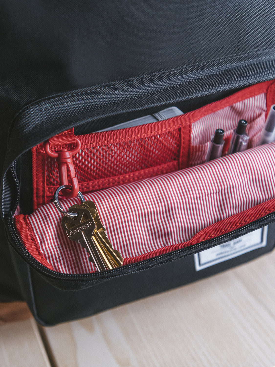 A close-up of an open Herschel Pop Quiz Backpack shows a red and white striped lining, keys on a red key hook, and pens in inner pockets—ideal for daily commutes. The Herschel pack is displayed on a light wooden surface.