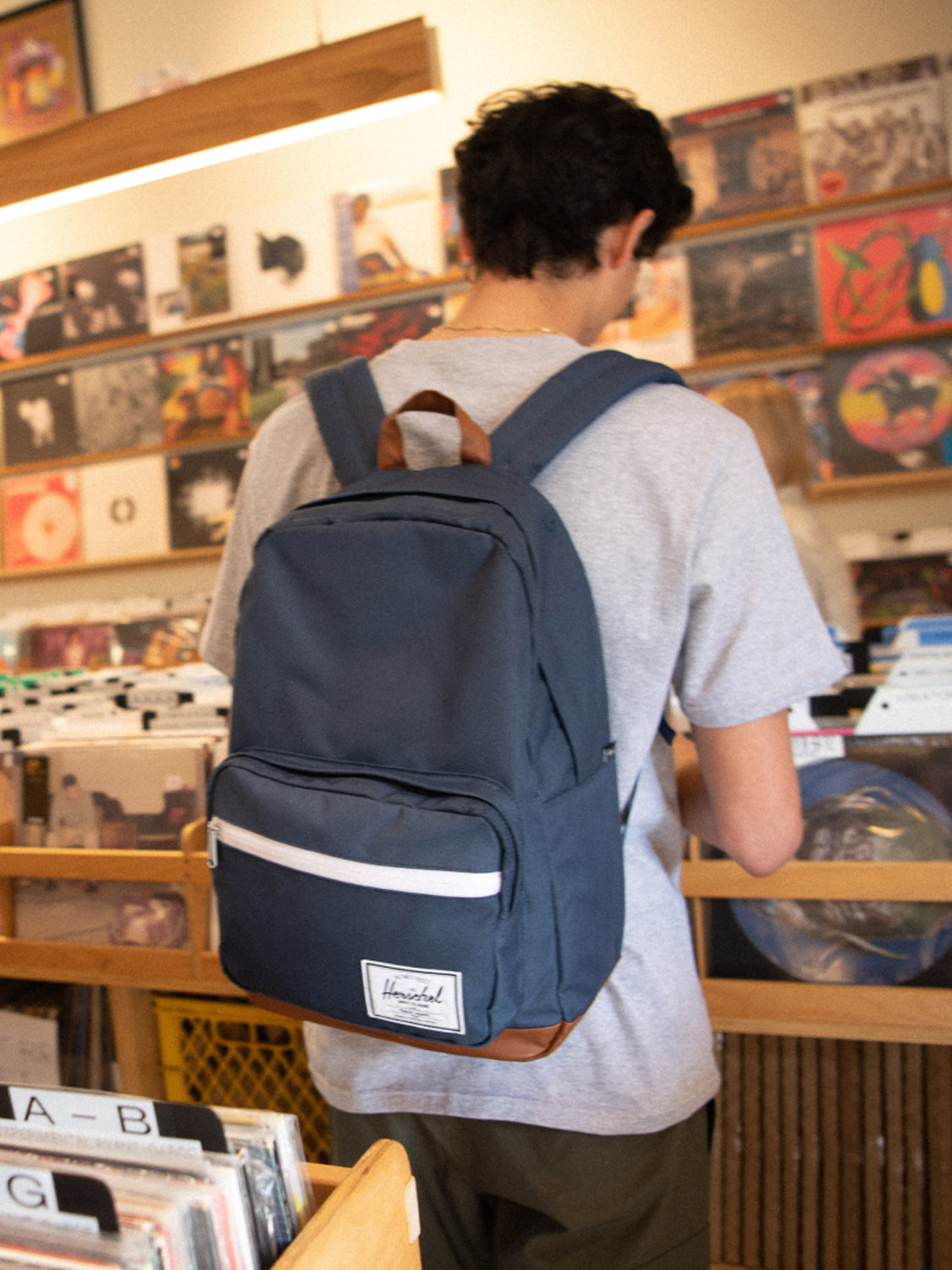A person with short dark hair, in a gray t-shirt and a navy blue Herschel Pop Quiz Backpack, browses vinyl records in a music store filled with wooden bins—perhaps taking a break from the daily commute.
