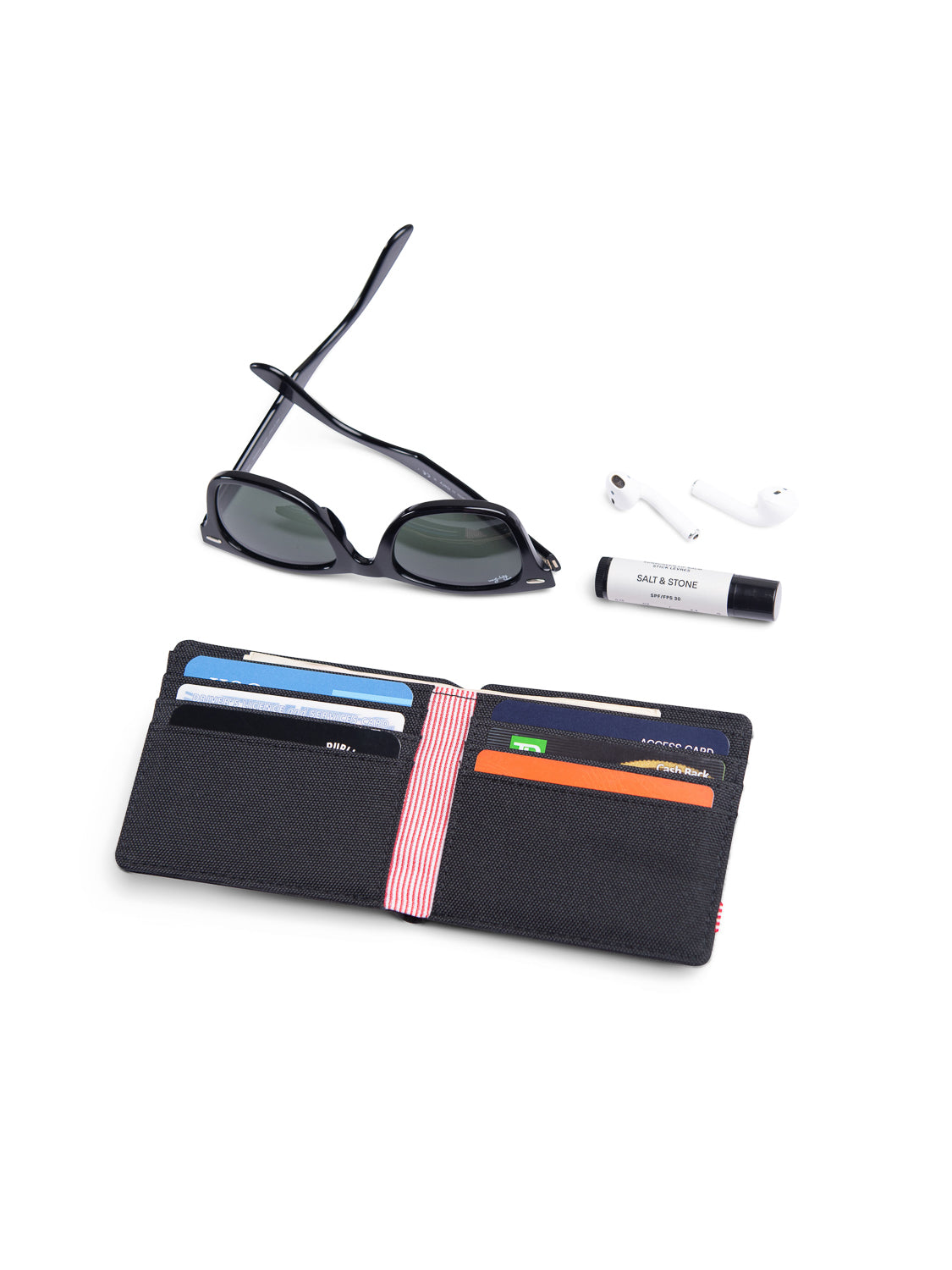 A pair of black sunglasses, white wireless earbuds, a tube of Salt & Stone lip balm, and an open Herschel Roy Classic Folded Wallet with multiple card slots are arranged on a white surface.