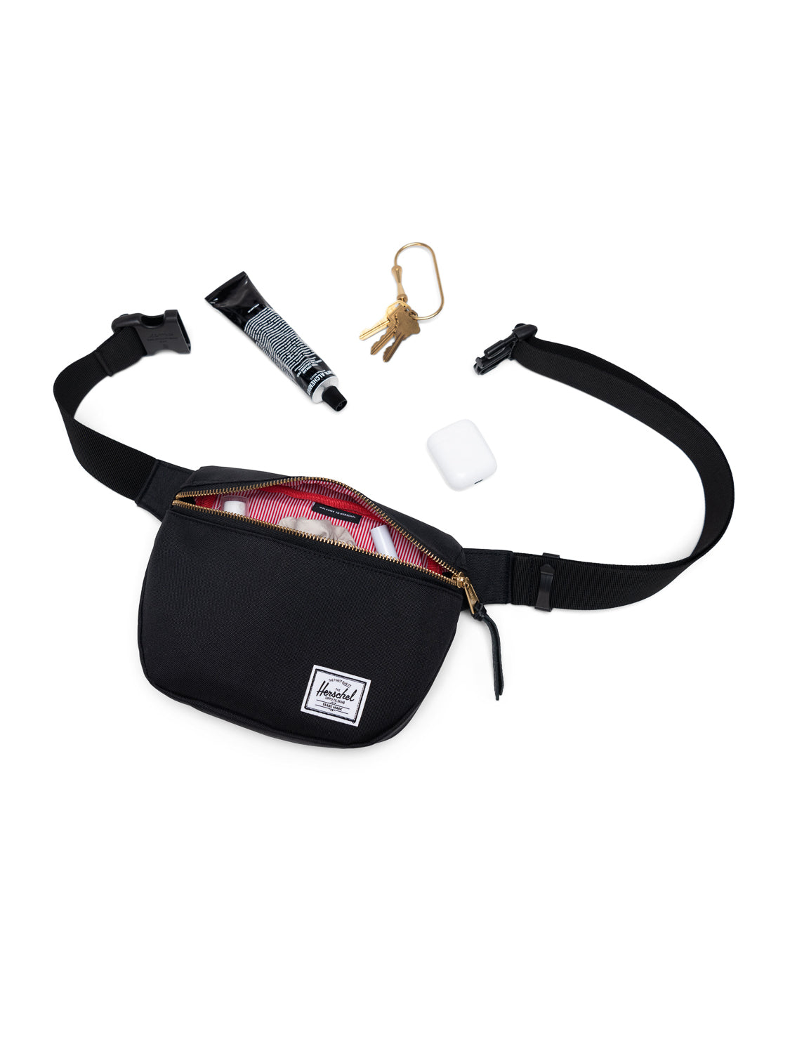 The black Herschel Fifteen Hip Pack with a brown metal zipper is open, showing a red lining. Nearby are hand cream, keys with a gold keyring, and a white wireless earbud case. The adjustable strap provides comfort on the go.