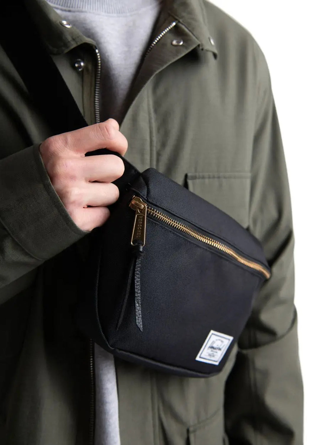 A person in an olive green jacket and gray shirt holds the adjustable strap of a Herschel Fifteen Hip Pack, a black crossbody bag featuring a metal zipper and a white logo patch.