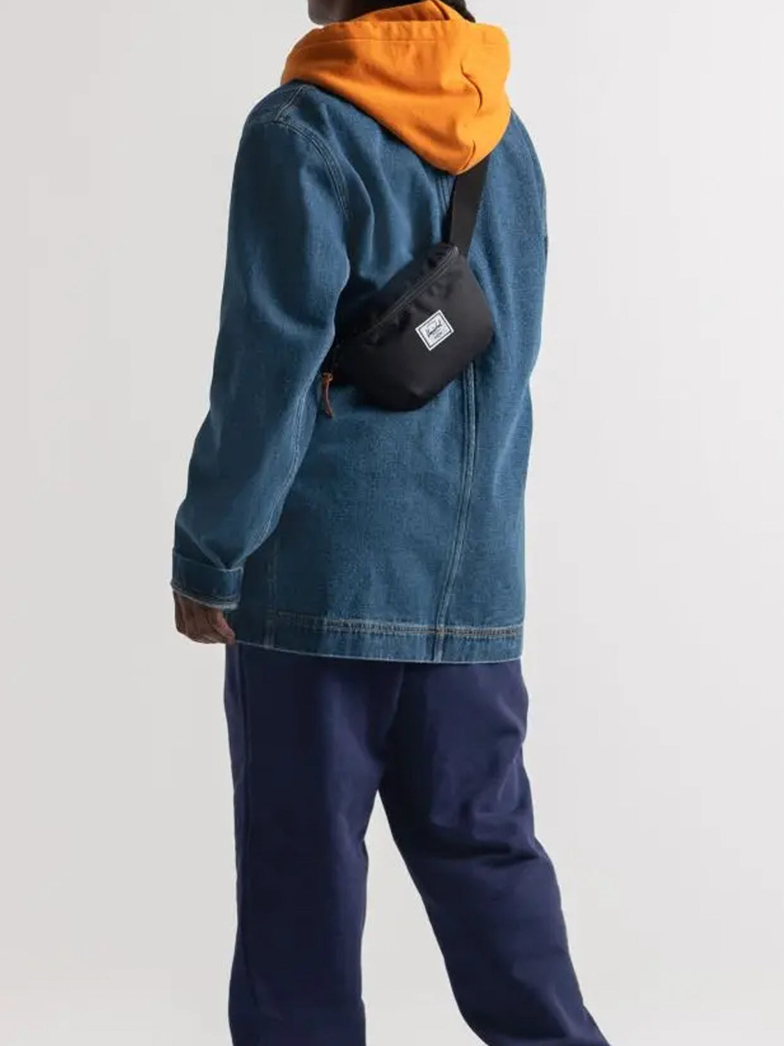 A person stands facing away, wearing a blue denim jacket, orange hoodie, navy pants, and a Herschel Fourteen Hip Pack with an adjustable strap and zippered main compartment against a plain light background.