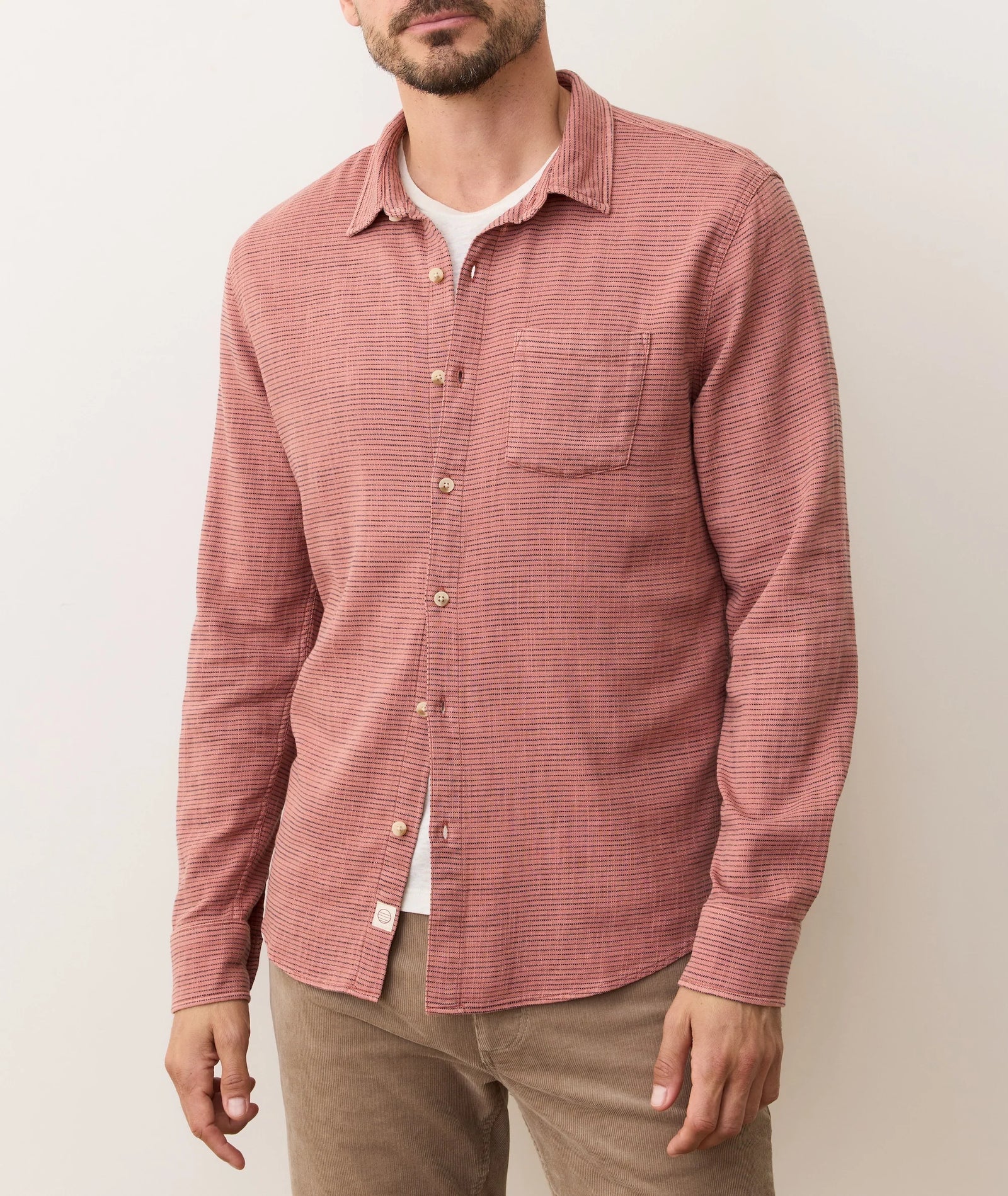 A man wears the Marine Layer Stretch Selvage Long Sleeve Shirt in red over a white tee and tan pants, standing against a plain light background with his face partially out of frame.