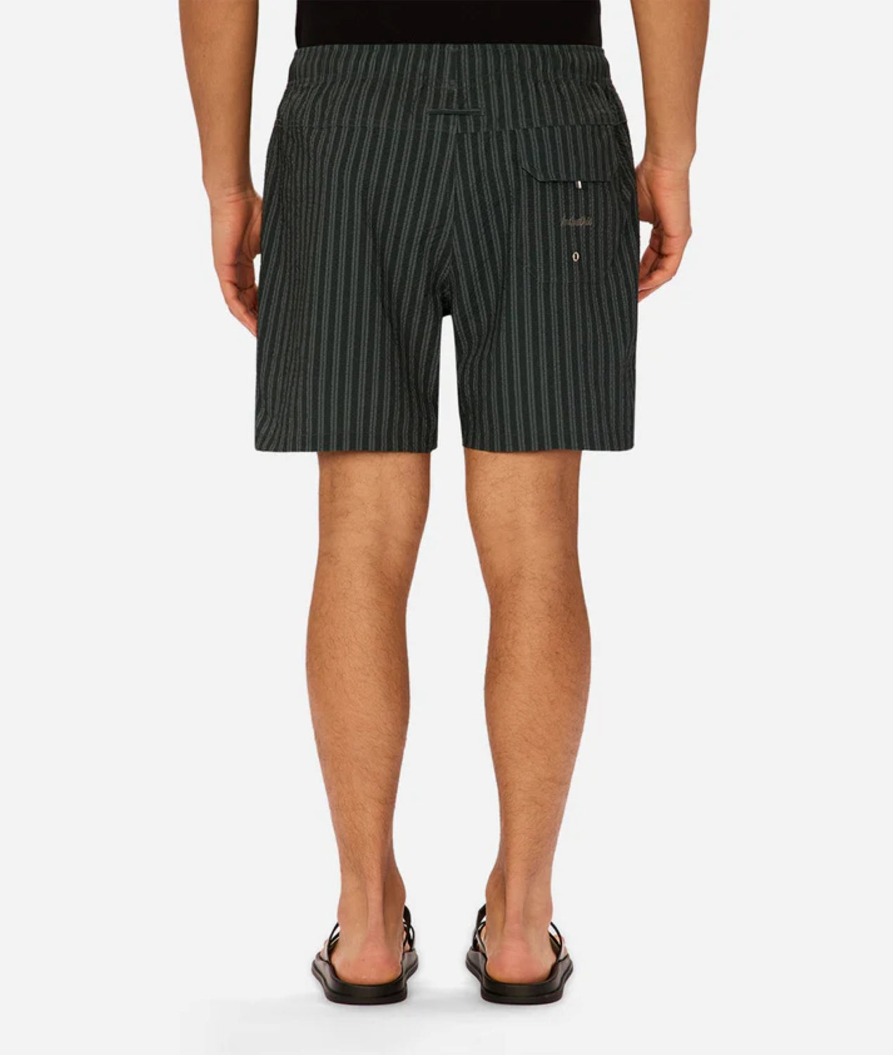 A person is seen from the back wearing Industrie’s Berkeley Swim Short in dark green and black pinstripes, paired with a black shirt and sandals. The shorts have a small rear button pocket.