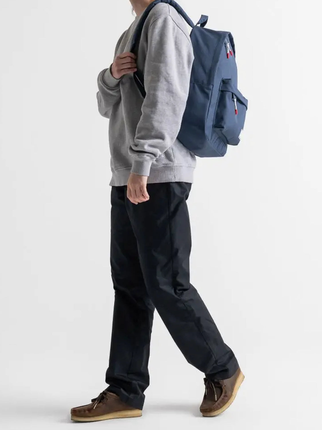 A person in a gray sweatshirt, black pants, and brown shoes walks with the Herschel Classic Backpack, ideal for daily use, slung over one shoulder against a plain white background.