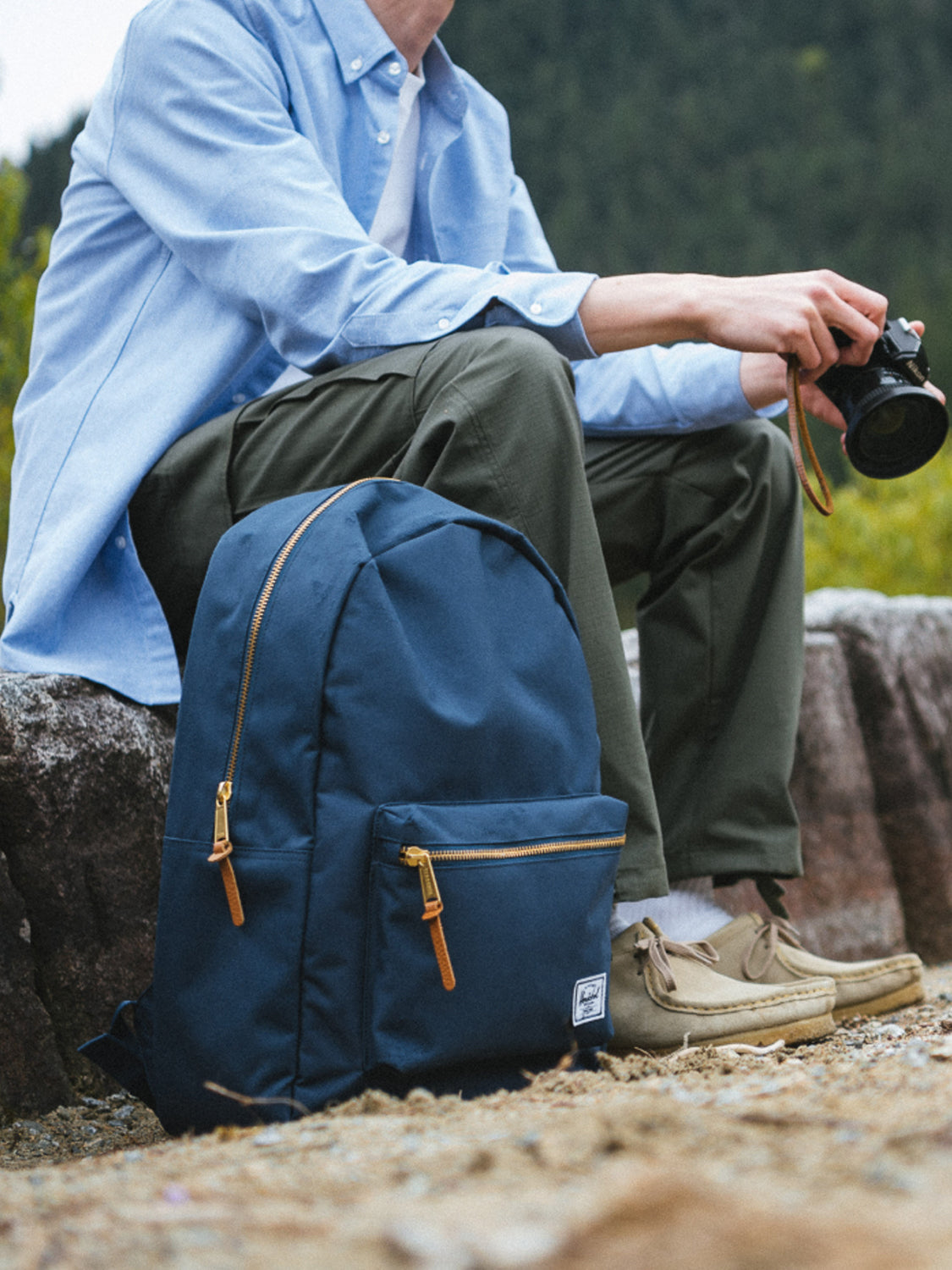 A person in a light blue shirt and olive pants sits on a stone ledge outdoors, holding a camera. Beside them is a Herschel Settlement Backpack with brown zippers. Trees and greenery create a tranquil backdrop.