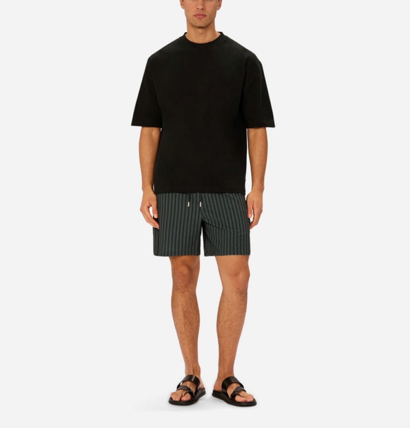 A man stands against a plain background, wearing Industrie’s black oversized T-shirt, green Berkeley Swim Short, and black sandals. His arms are relaxed at his sides and his expression is neutral.