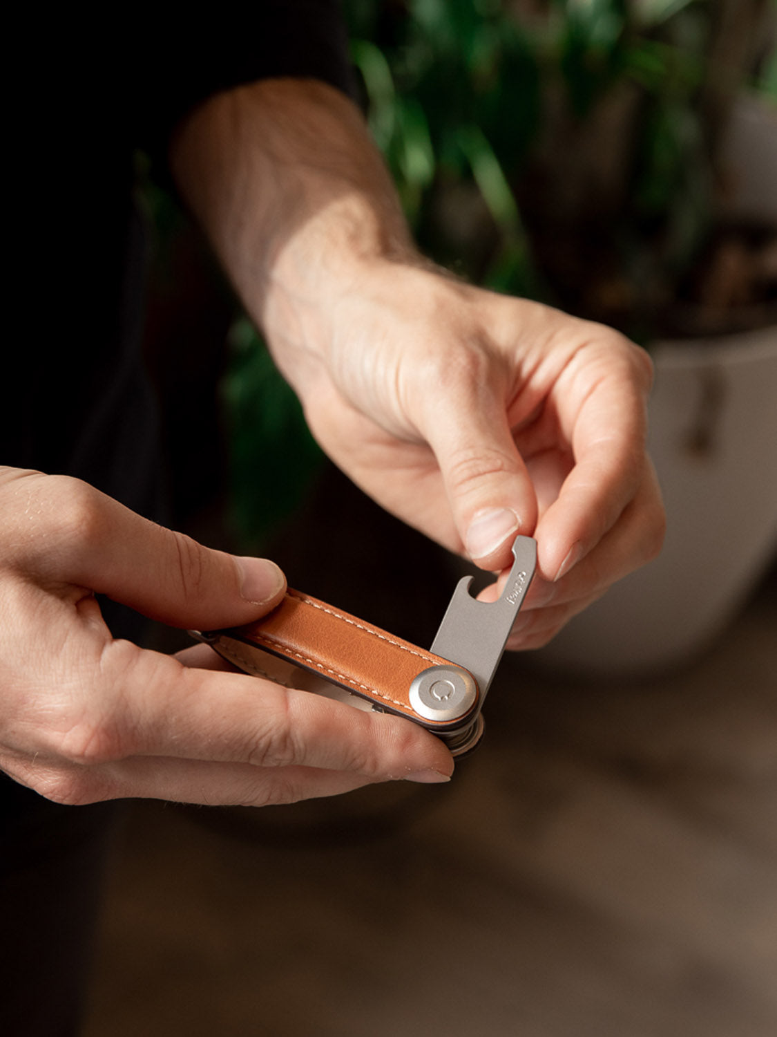 Someone holds an Orbitkeys stainless steel bottle opener, partly unfolded, featuring a brown leather grip. The background is softly blurred with hints of green plant leaves.