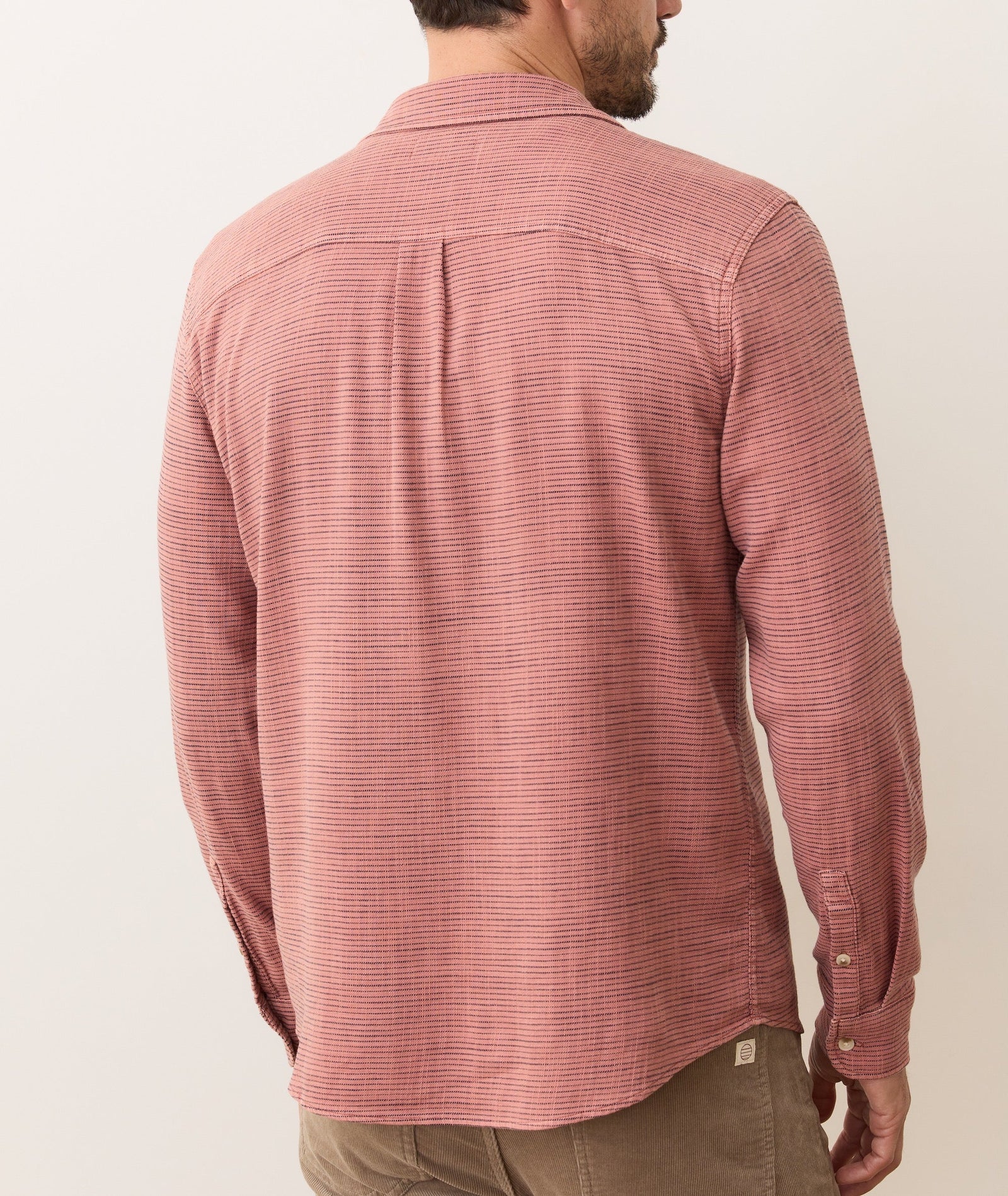 A man with short hair and a trimmed beard is seen from behind wearing a Marine Layer Stretch Selvage Long Sleeve Shirt in red and white stripes with brown pants; the shirt features a subtle textured pattern.