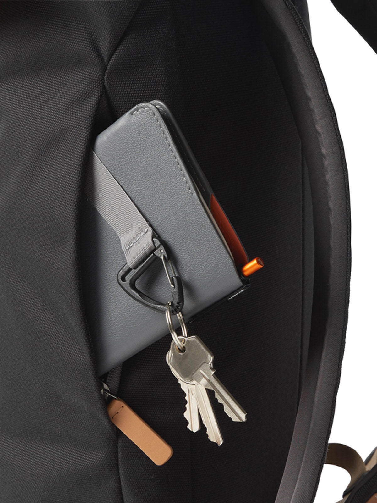A close-up of a gray key holder clipped to the pocket of a Bellroy Oslo Backpack - 16L, carrying several silver keys and revealing part of a small orange object inside the slim backpack pocket.
