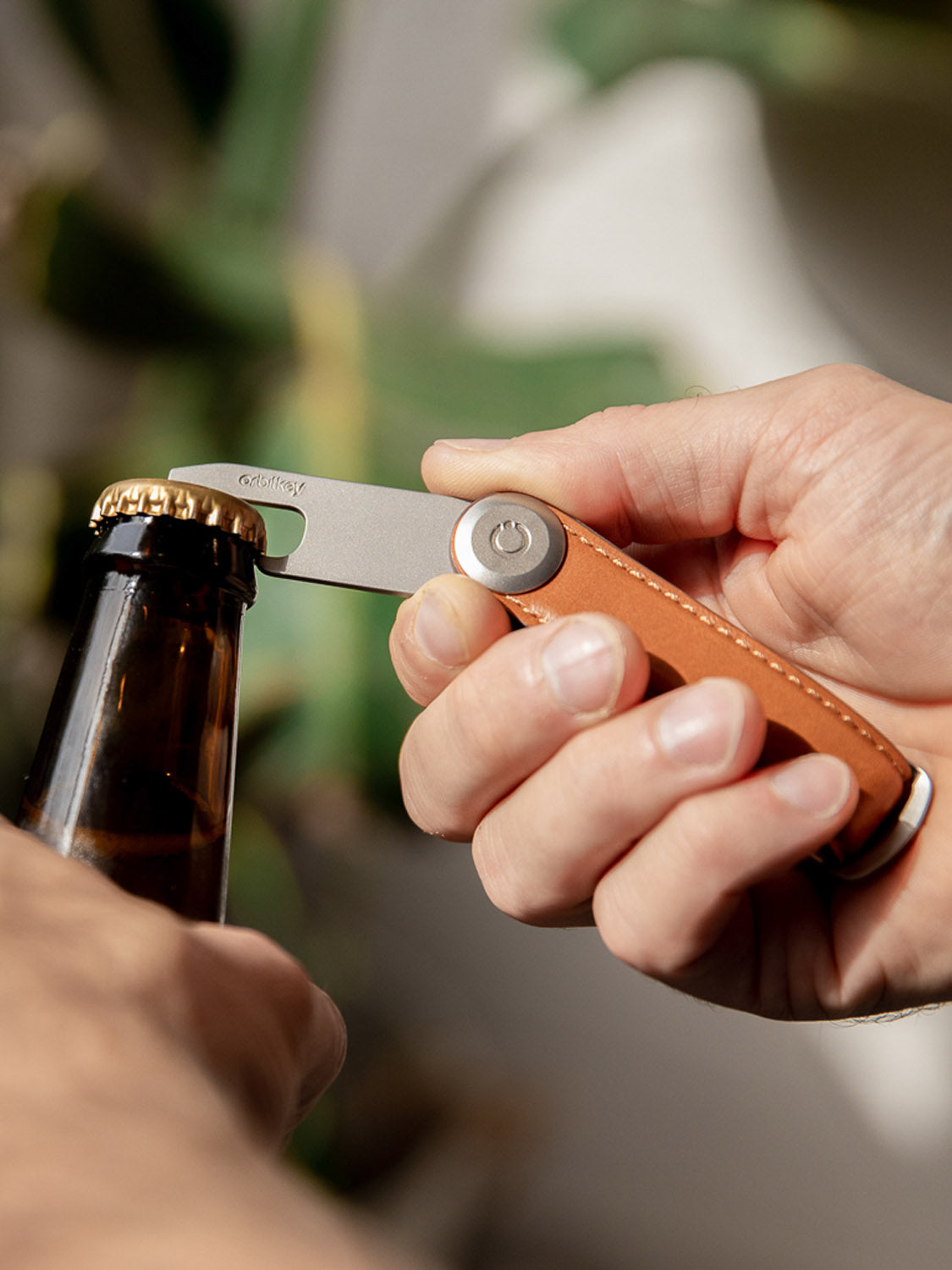 A hand uses the Orbitkey Stainless Steel Bottle Opener to pry a cap from a dark bottle. The multitool boasts a slim design, complemented by a brown leather handle, while blurred green leaves in the background enhance the natural charm of the scene.