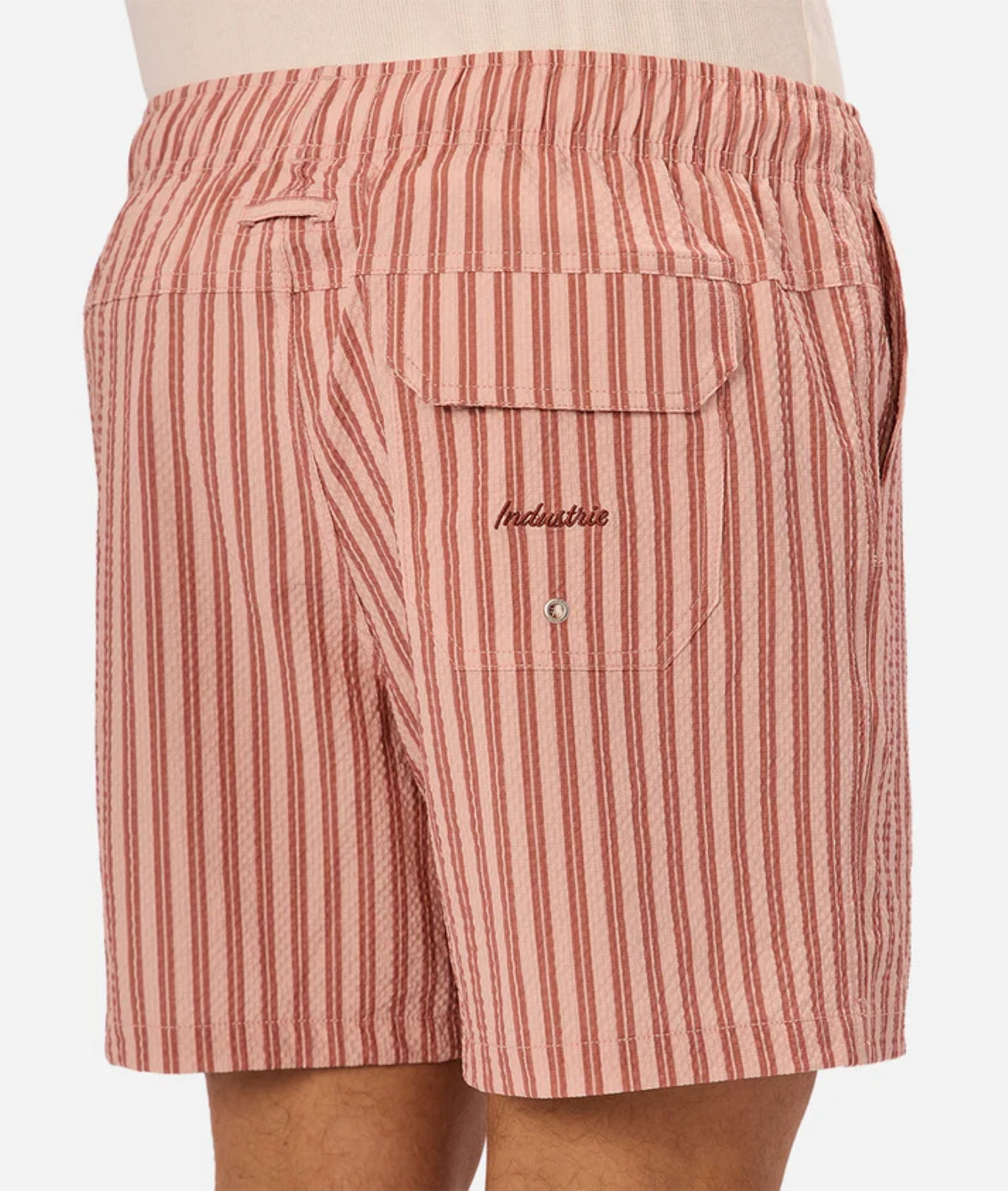 A close-up of a person wearing Industrie’s Berkeley Swim Short—red and white vertical seersucker stripes, elastic waistband, and a button-flap back pocket with Industrie embroidery above it.
