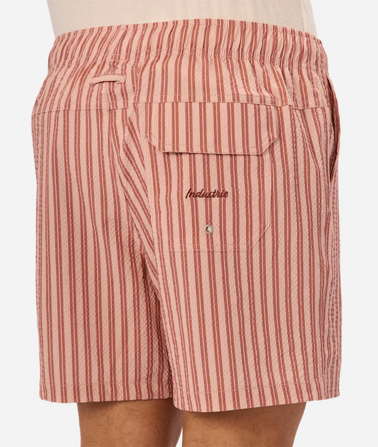 A close-up of a person wearing Industrie’s Berkeley Swim Short—red and white vertical seersucker stripes, elastic waistband, and a button-flap back pocket with Industrie embroidery above it.