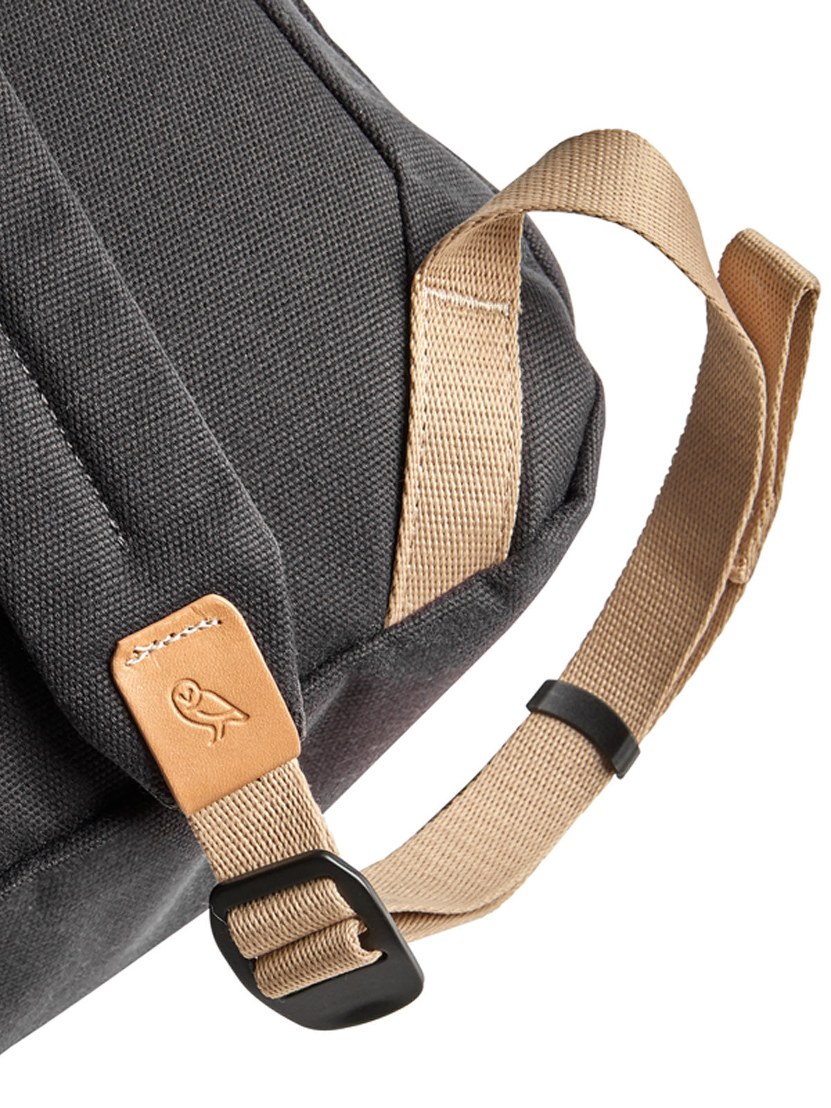Close-up of the Bellroy Oslo Backpack - 16L in dark gray with tan adjustable straps, a black plastic buckle, and a tan leather accent embossed with an owl logo and white text. Set against a white background.