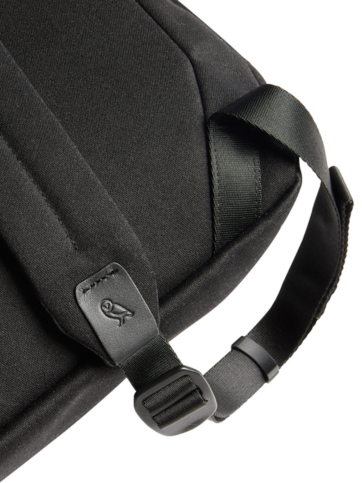 Close-up of the Bellroy Oslo Backpack - 16L in black fabric, featuring a strap with buckle and a small metal owl logo tag near the strap, set against a plain white background.