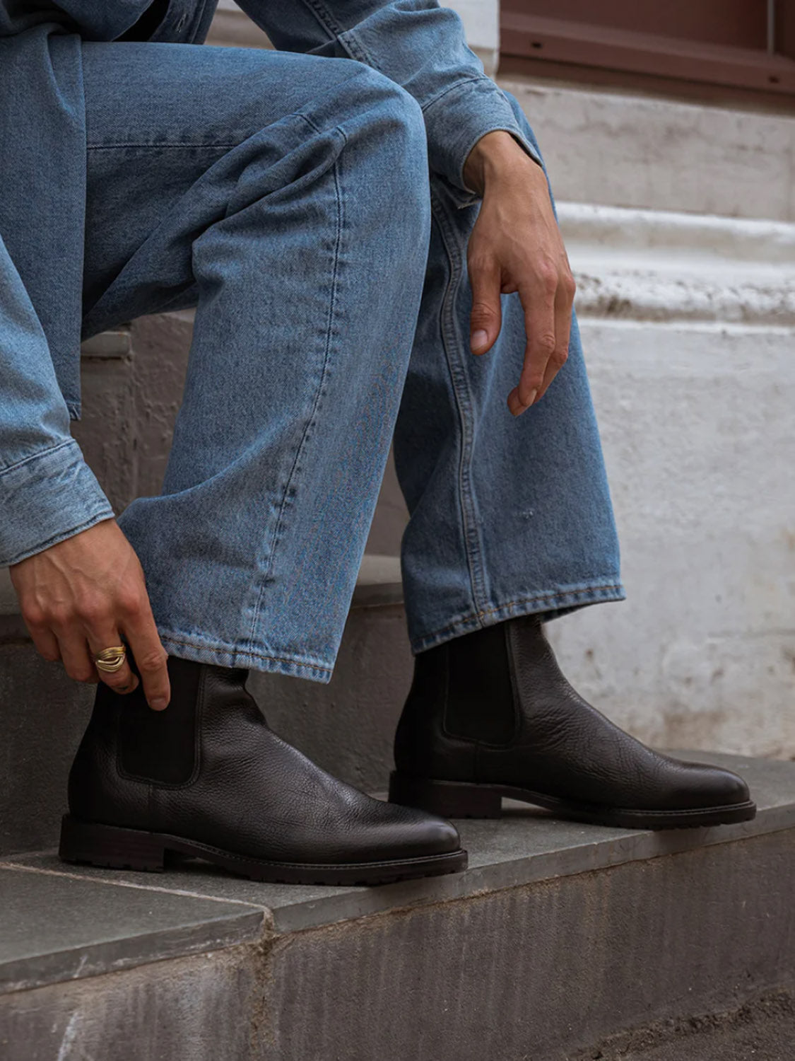 Seated on light stone steps, a person in light blue jeans and a denim shirt adjusts a sleek York Classic Chelsea Boot from Shoe The Bear. Their relaxed posture reveals a gold ring on their hand, with the flexible rubber outsole of the boot enhancing its elegance.
