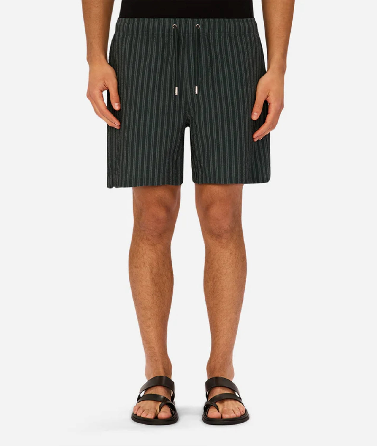 A person wearing Industrie’s Berkeley Swim Short in dark green pinstripe, paired with a black top and black sandals, standing against a plain white background. Image is cropped from shoulders down.