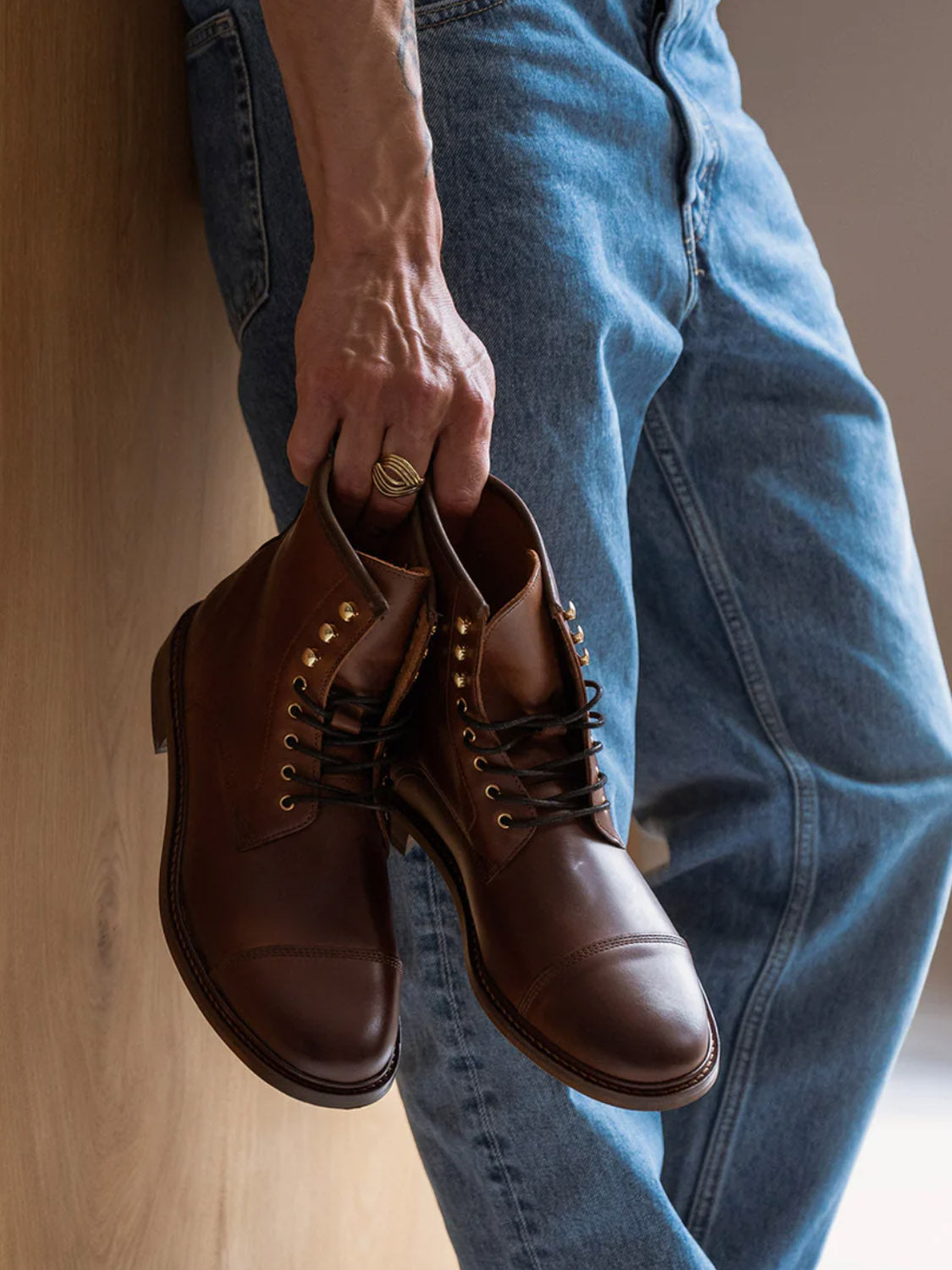 A person in blue jeans stands against a wooden wall, holding the Curtis Hand Crafted Lace-Up Leather Boots by Shoe The Bear. Made from LWG-certified leather with gold eyelets, these boots exude elegance. A gold ring adorns one finger, enhancing the stylish yet casual ambiance.