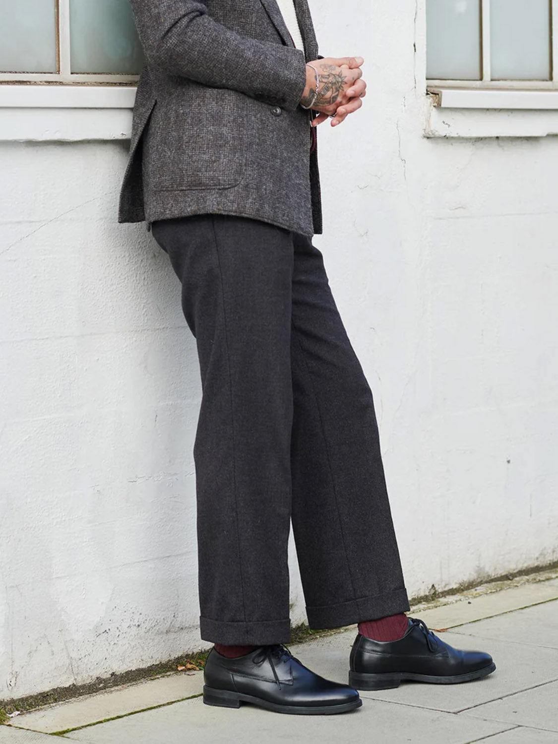 A person in a gray suit stands against a white wall, hands clasped. They wear cropped trousers, striking red socks, and Shoe The Bears Linea Derby Style shoes made of LWG-certified leather. Their face is hidden, displaying only a tattoo on the hand. The scene exudes an urban vibe.