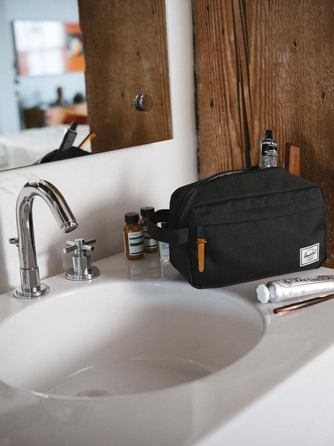 The Herschel Chapter Travel Kit, featuring a waterproof zipper, rests on a white bathroom sink next to a faucet, small bottles, a toothpaste tube, and a toothbrush, with a wooden mirror frame and a softly blurred background.