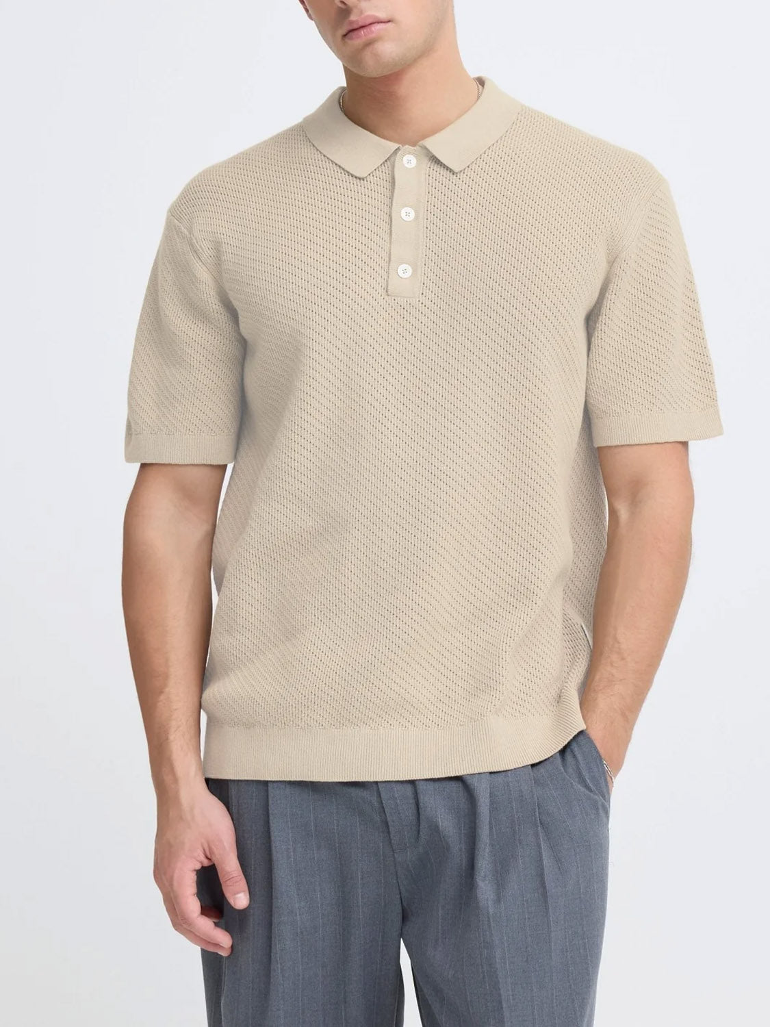 A man wears the Cfisaksson Polo Knit by Casual Friday—100% cotton, beige, with a textured pattern—paired with gray pleated trousers. He stands with one hand in his pocket against a plain background, perfect for a smart Casual Friday look.