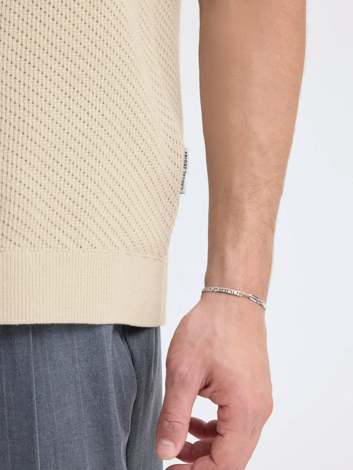 A close-up of a person's right arm wearing the Casual Friday Cfisaksson Polo Knit, a light beige textured 100% cotton sweater, paired with grey pinstripe pants and a thin silver chain bracelet. The background is plain and light-colored.